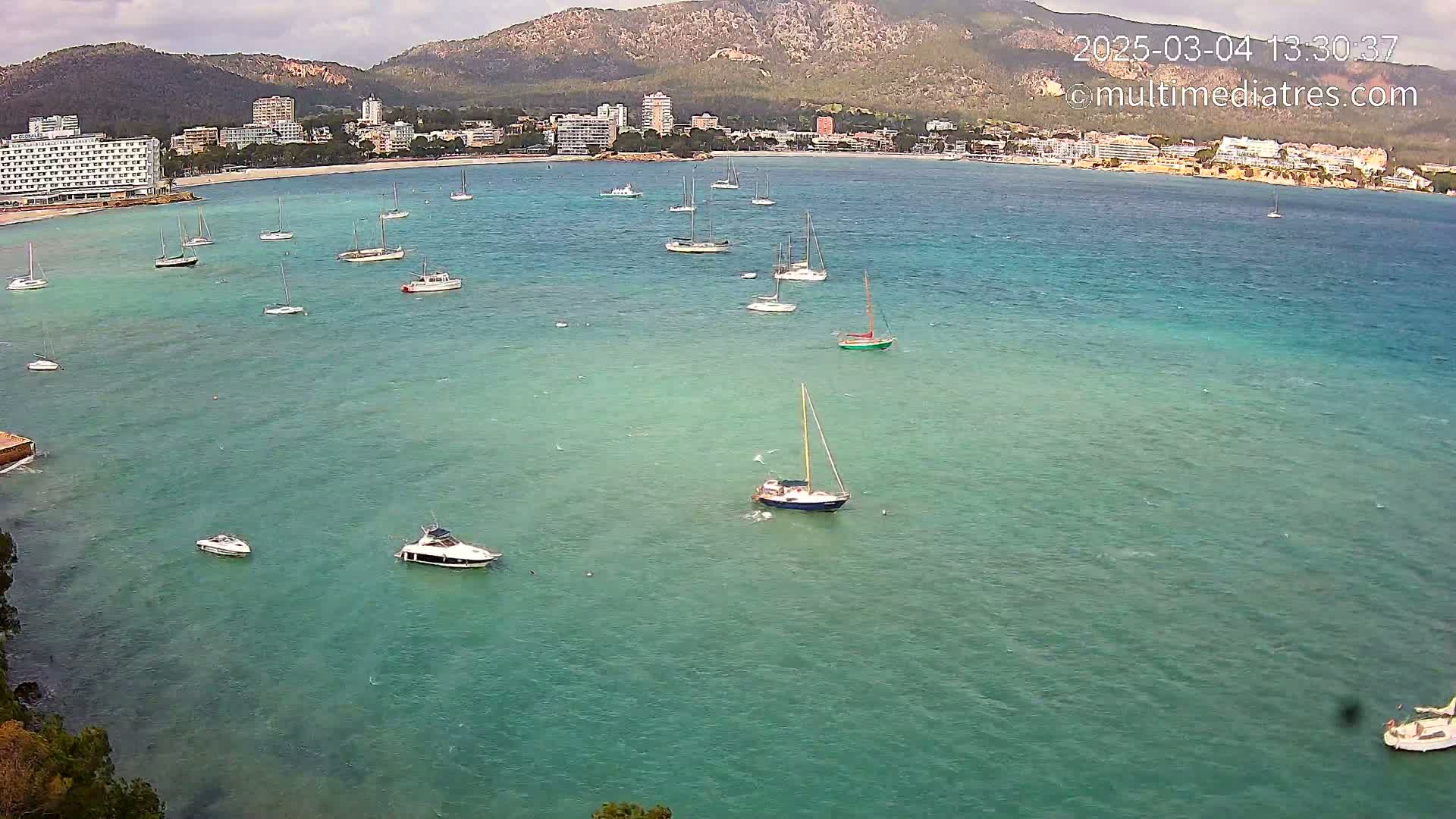 Numerous sailboats and motorboats are anchored in a calm, teal-colored bay, with a shoreline of hotels and buildings visible in the background under a partly cloudy sky.