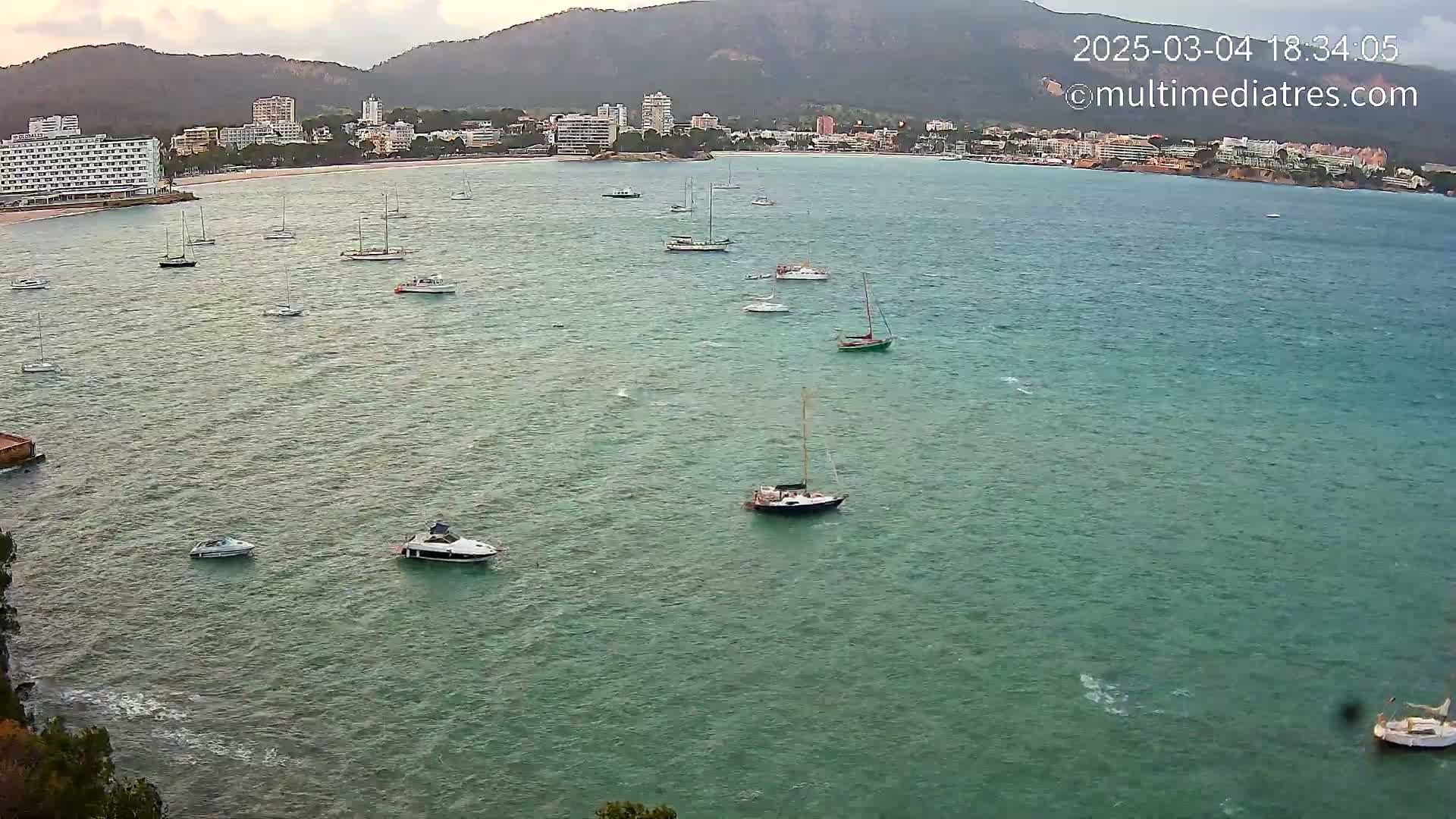 Numerous sailboats and motorboats are anchored in a calm, teal-colored bay, with a town and mountains visible in the background under a partly cloudy sky.