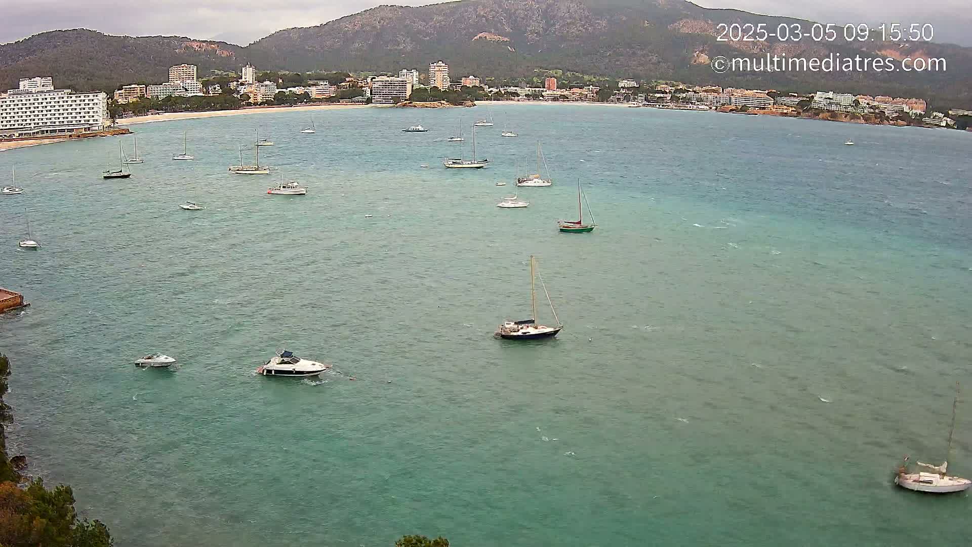 Numerous boats are anchored in a calm, turquoise bay, with a shoreline of buildings and a mountainous backdrop under an overcast sky.