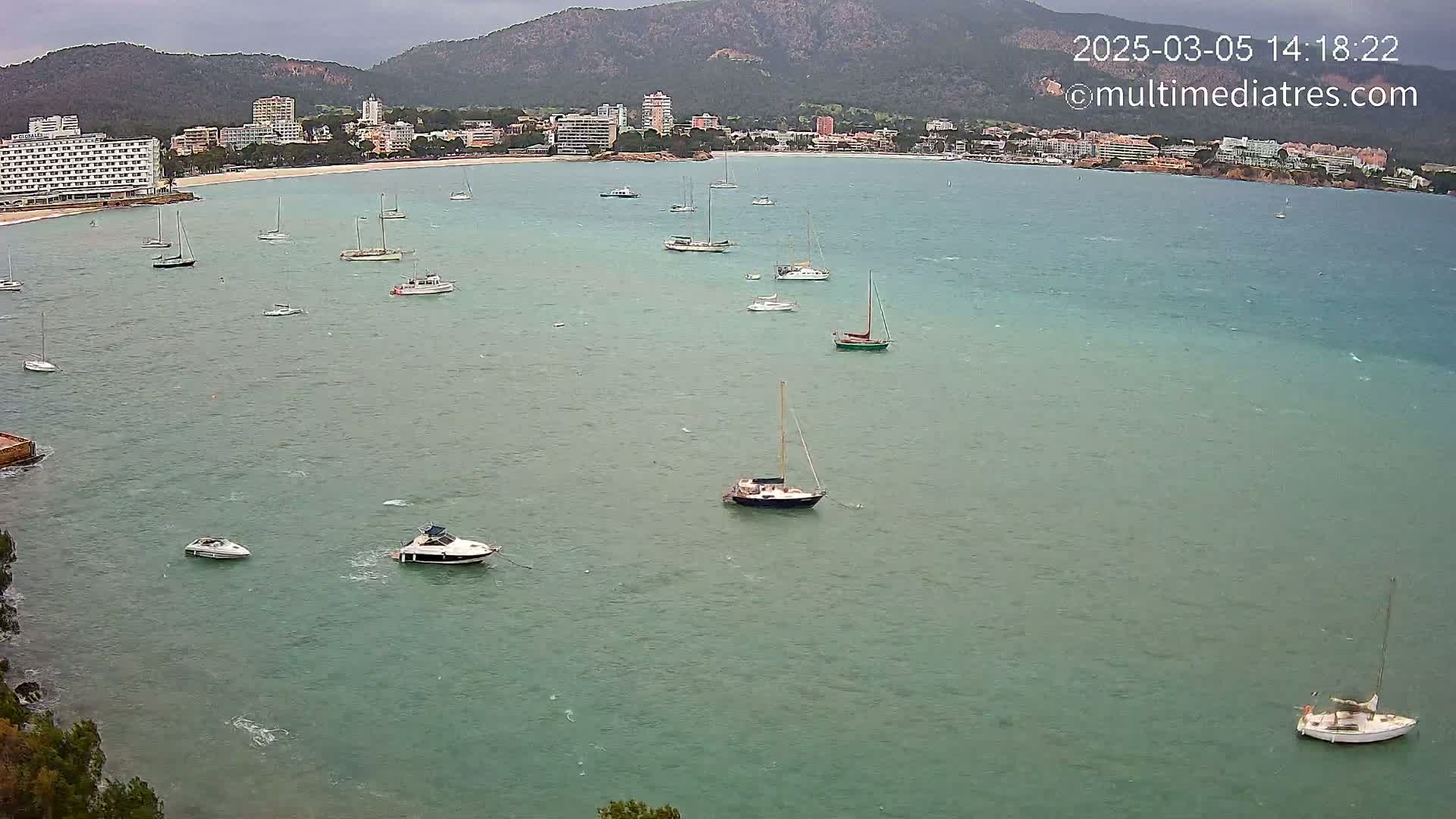 Numerous sailboats and motorboats are scattered across a calm, green-blue bay under an overcast sky, with a coastline of hotels and buildings visible in the distance.