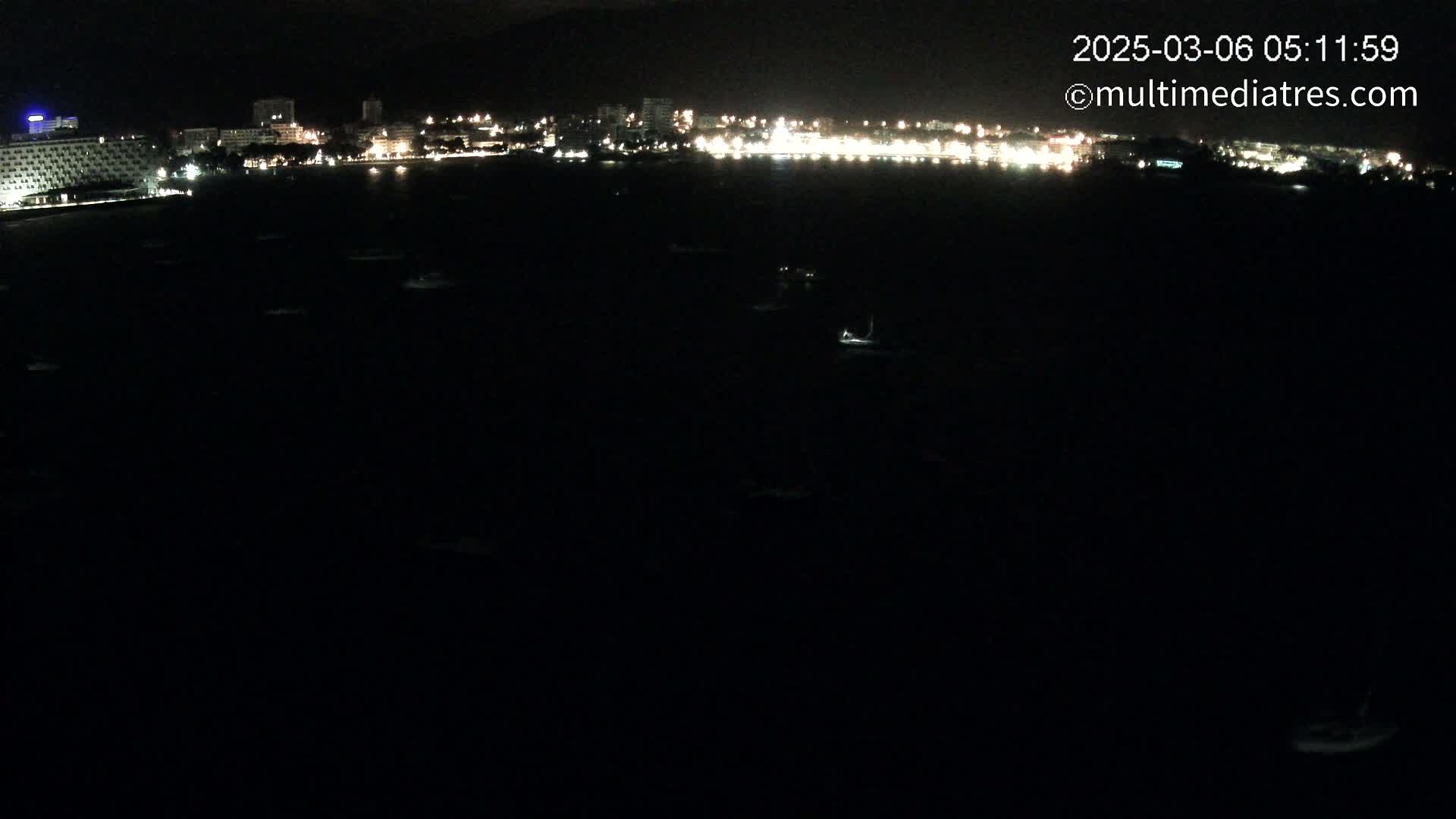 A dark body of water at night displays a string of lights from a distant city on the horizon, with a few small lights scattered on the water.
