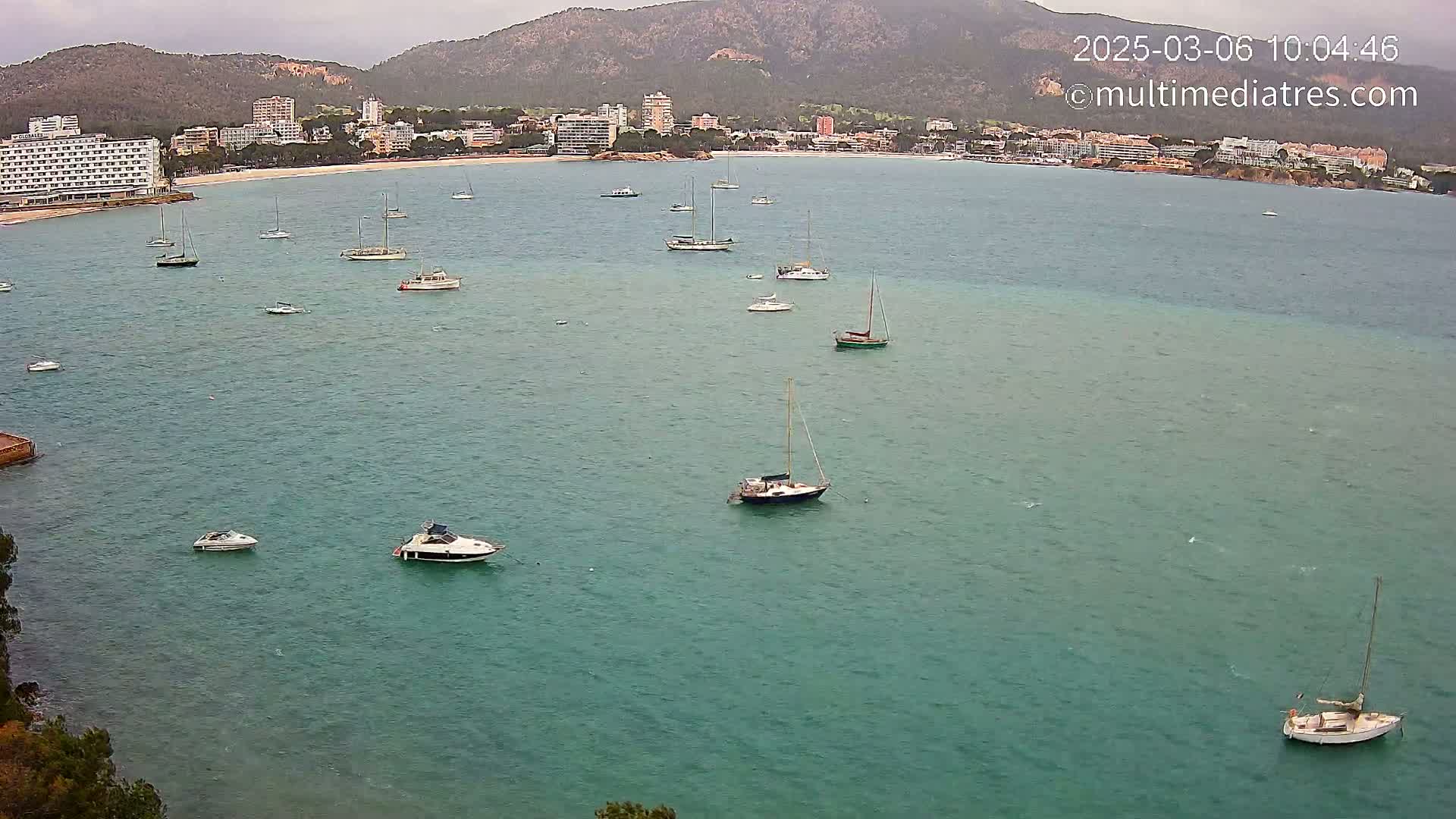 Numerous sailboats and motorboats are anchored in a calm, teal bay near a shoreline with hotels and mountains in the background under an overcast sky.