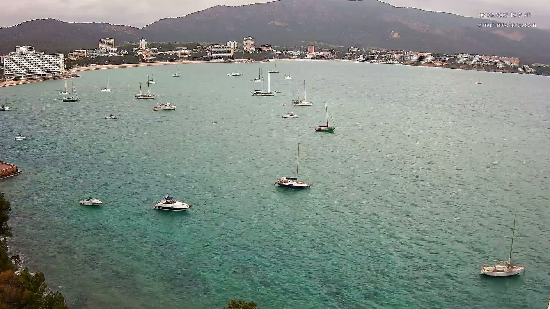 A calm, teal-colored bay with numerous sailboats and motorboats anchored near a shoreline featuring buildings and hills under an overcast sky.