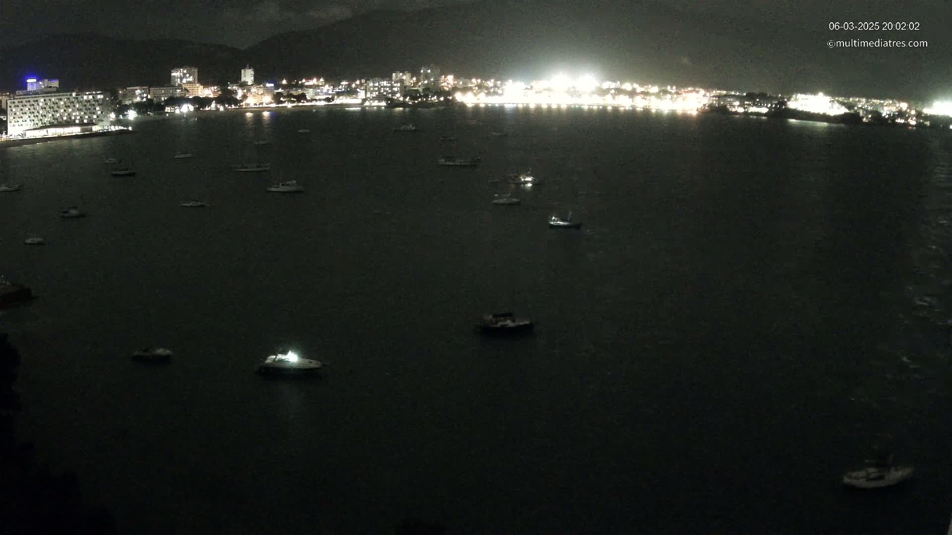Numerous boats are moored in a dark body of water at night, in front of a brightly lit city skyline.