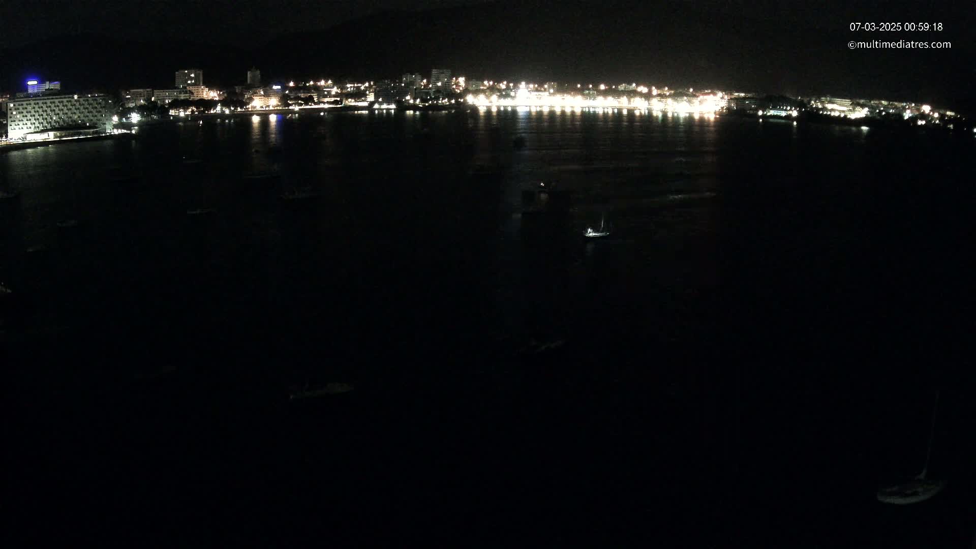 A nighttime, dark view of a city's illuminated waterfront at night, with several boats in the dark water.