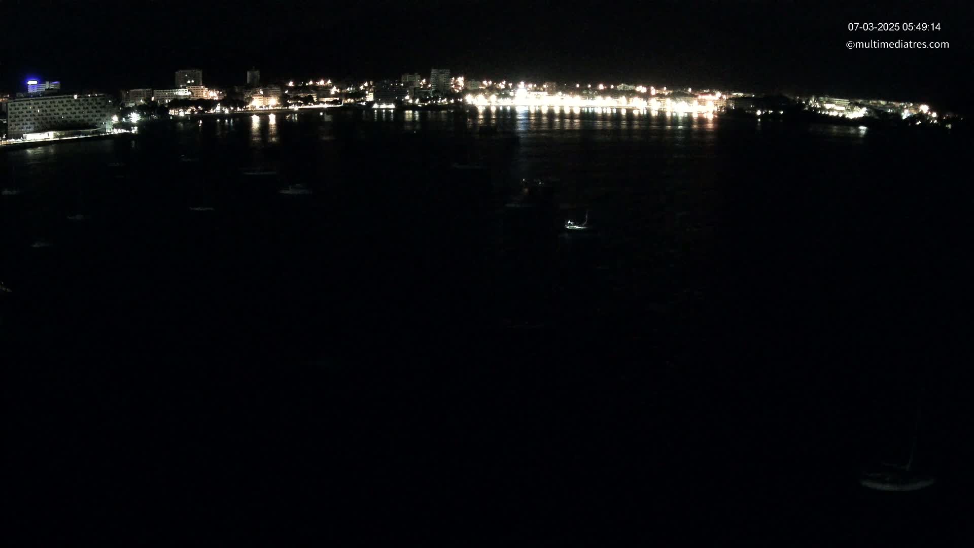 A nighttime view of a city's illuminated waterfront, with several small boats scattered across a dark body of water.