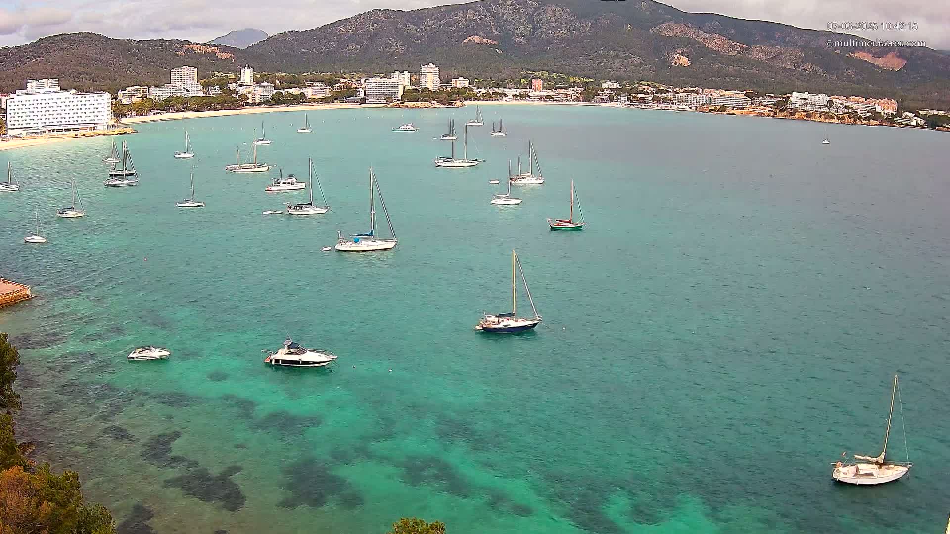 A calm, turquoise bay dotted with numerous sailboats and motorboats, sits before a coastal town nestled against a mountain range under an overcast sky.