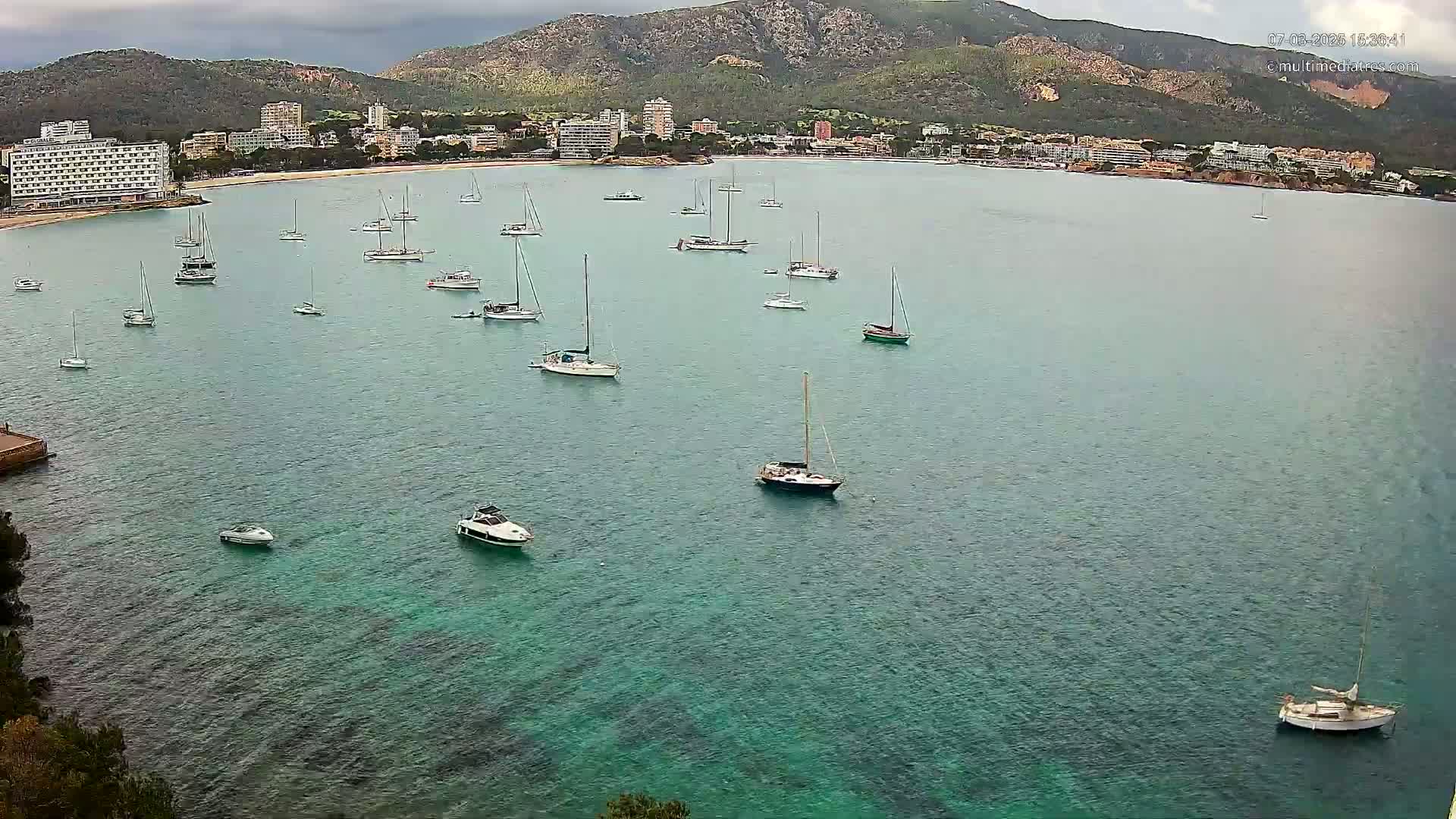 Numerous sailboats and motorboats are anchored in a calm, teal bay, with a backdrop of a mountainous coastline and buildings under an overcast sky.