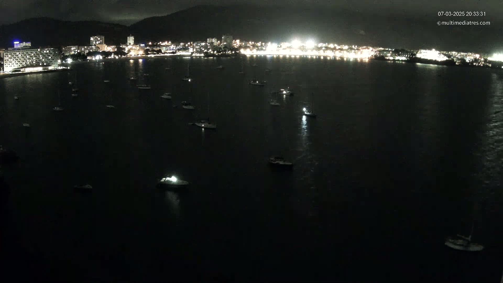 At night, numerous boats are moored in a dark body of water in front of a brightly lit city nestled against a mountain.