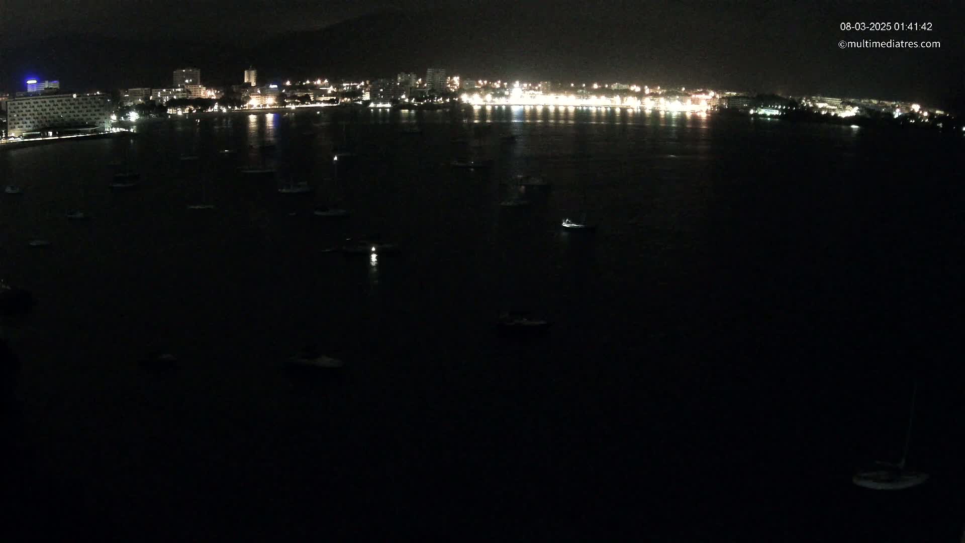 A dark night scene shows a city's illuminated waterfront at night with numerous small boats scattered across a dark body of water.