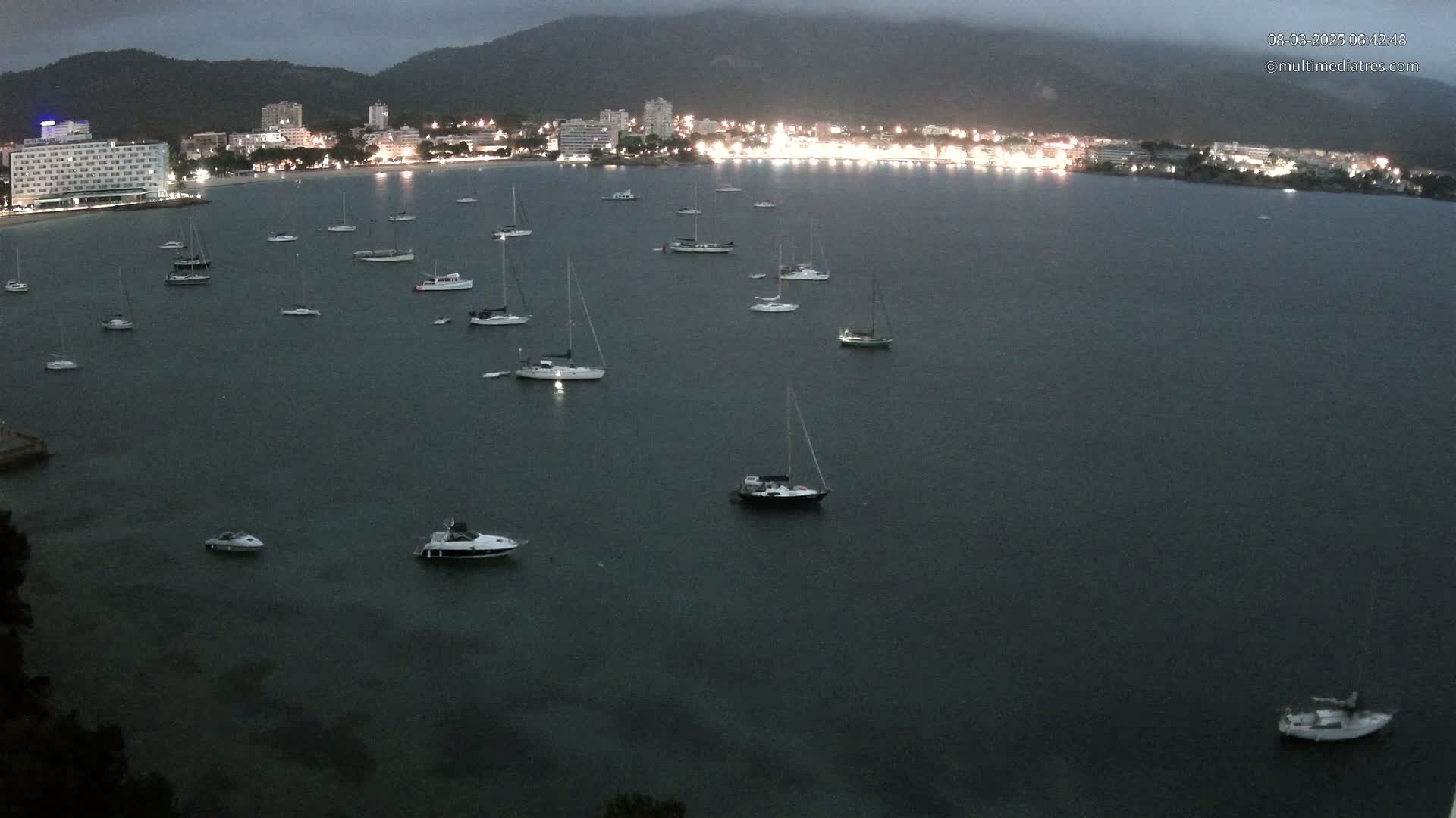 Many sailboats and motorboats are moored in a calm, dark bay at night, with a city's lights illuminating the shoreline in the distance.