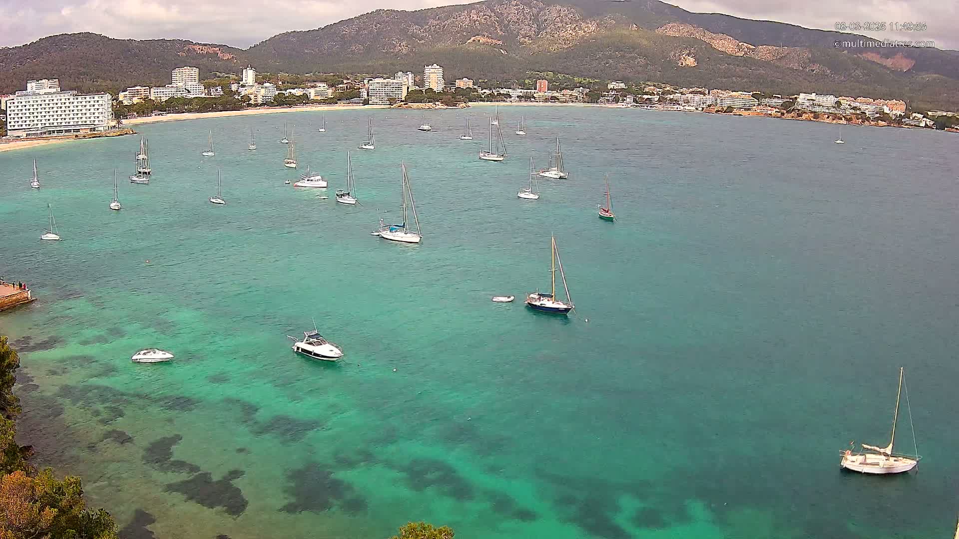 Numerous sailboats and motorboats are anchored in a calm, clear turquoise bay near a coastline with hotels and mountains under a cloudy sky.