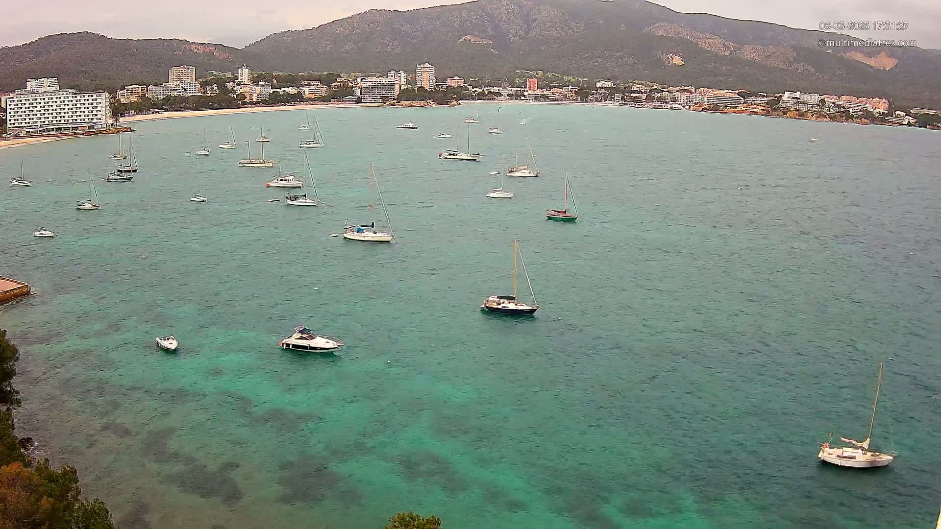 Numerous sailboats and motorboats are anchored in a calm, teal-colored bay near a shoreline with buildings and a mountain range under an overcast sky.