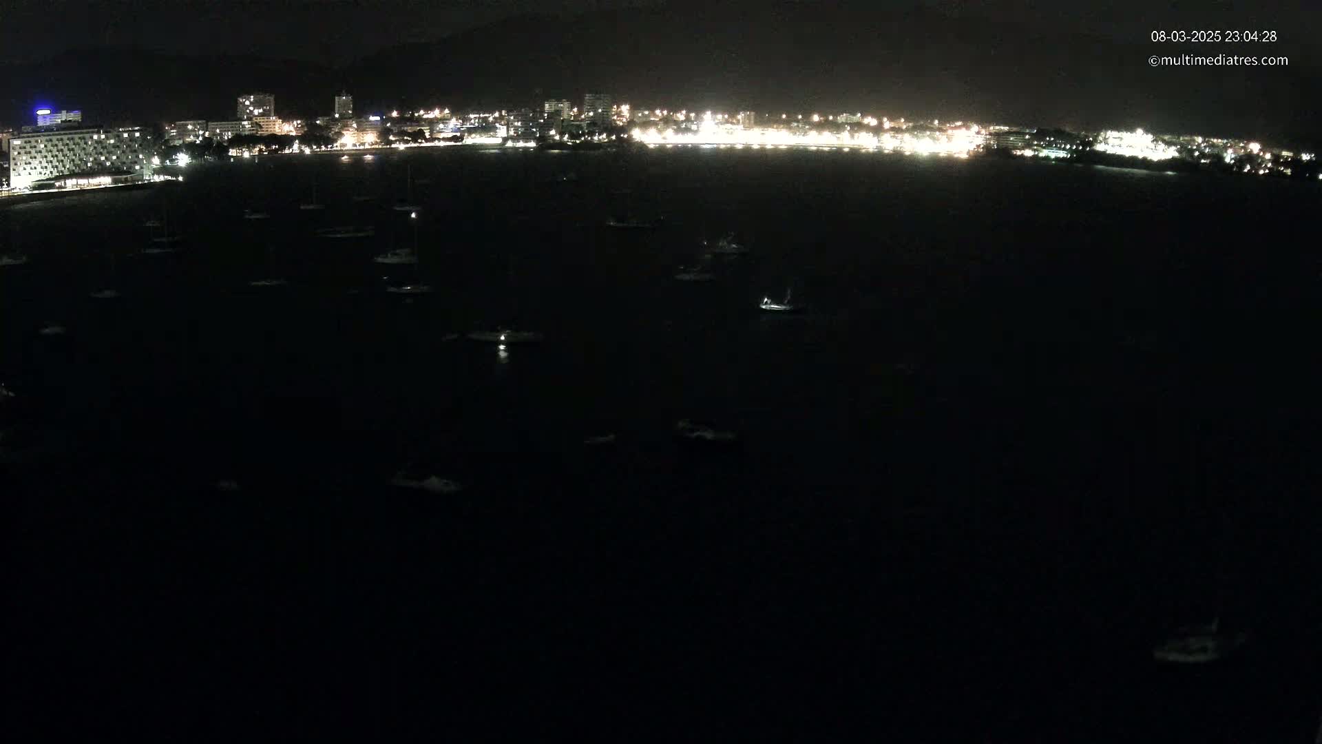 At night, numerous small boats are scattered across a dark body of water in front of a brightly lit city skyline.