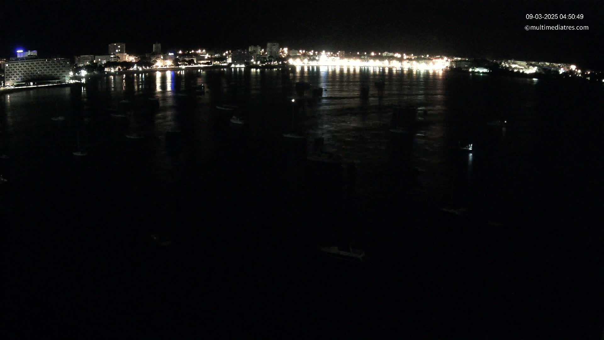At night, a dark body of water is speckled with numerous small boats near a brightly lit shoreline.