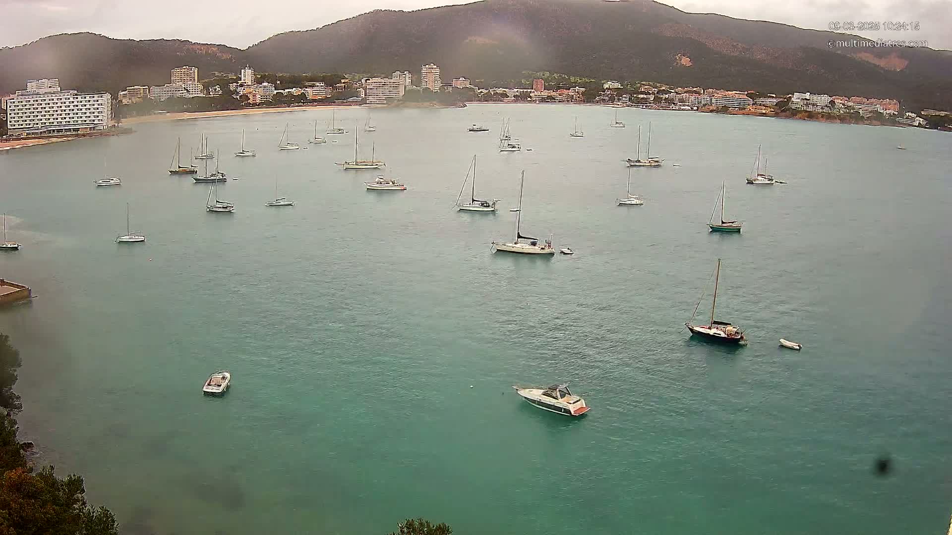 Numerous sailboats are anchored in a calm, teal-colored bay under an overcast sky near a shoreline with hotels and buildings.