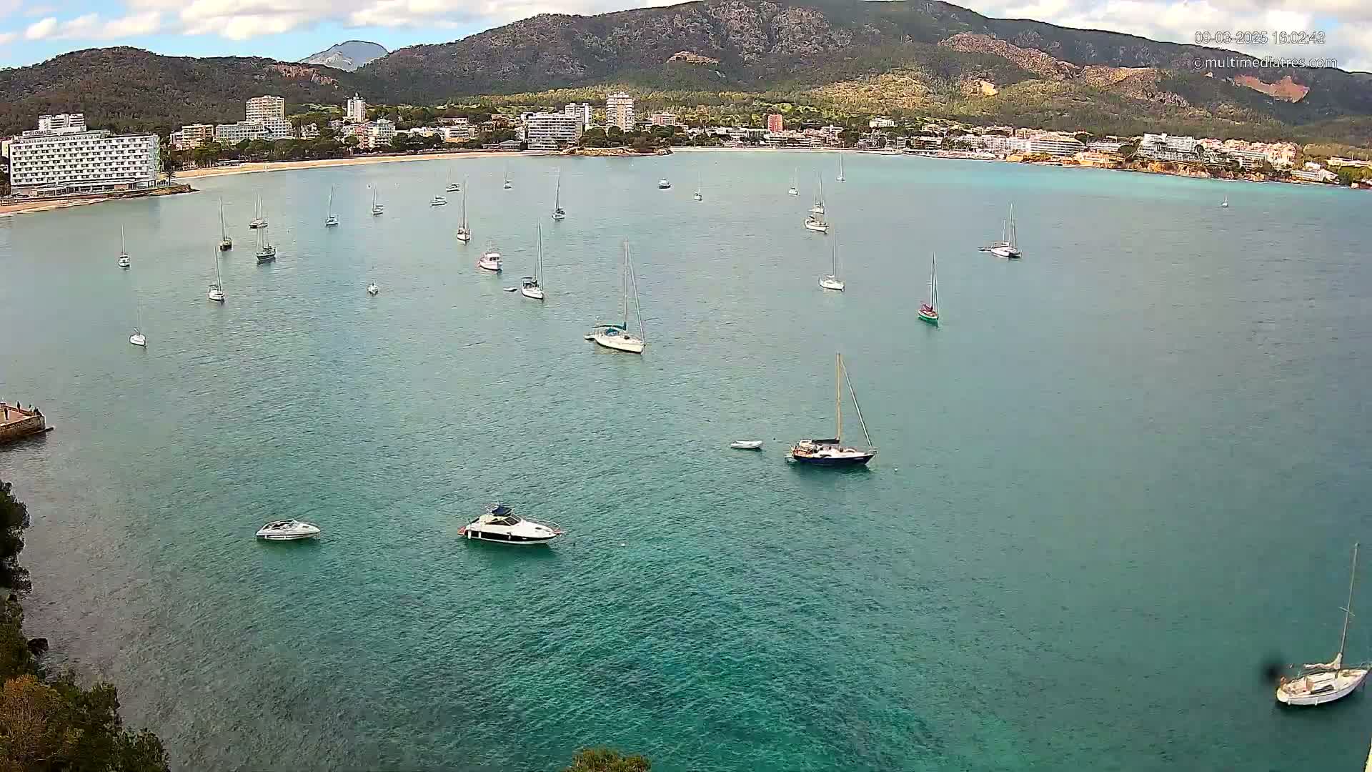 A calm, turquoise bay with numerous sailboats and motorboats is seen from an elevated vantage point, with a mountainous backdrop under partly cloudy skies.