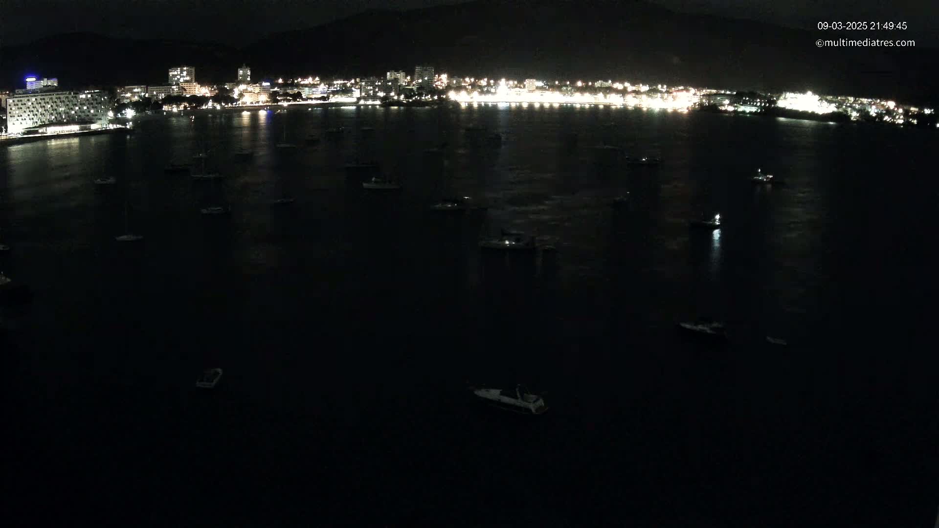 At night, a dark body of water dotted with numerous small boats is seen in front of a brightly lit coastal city situated against a dark mountain backdrop.