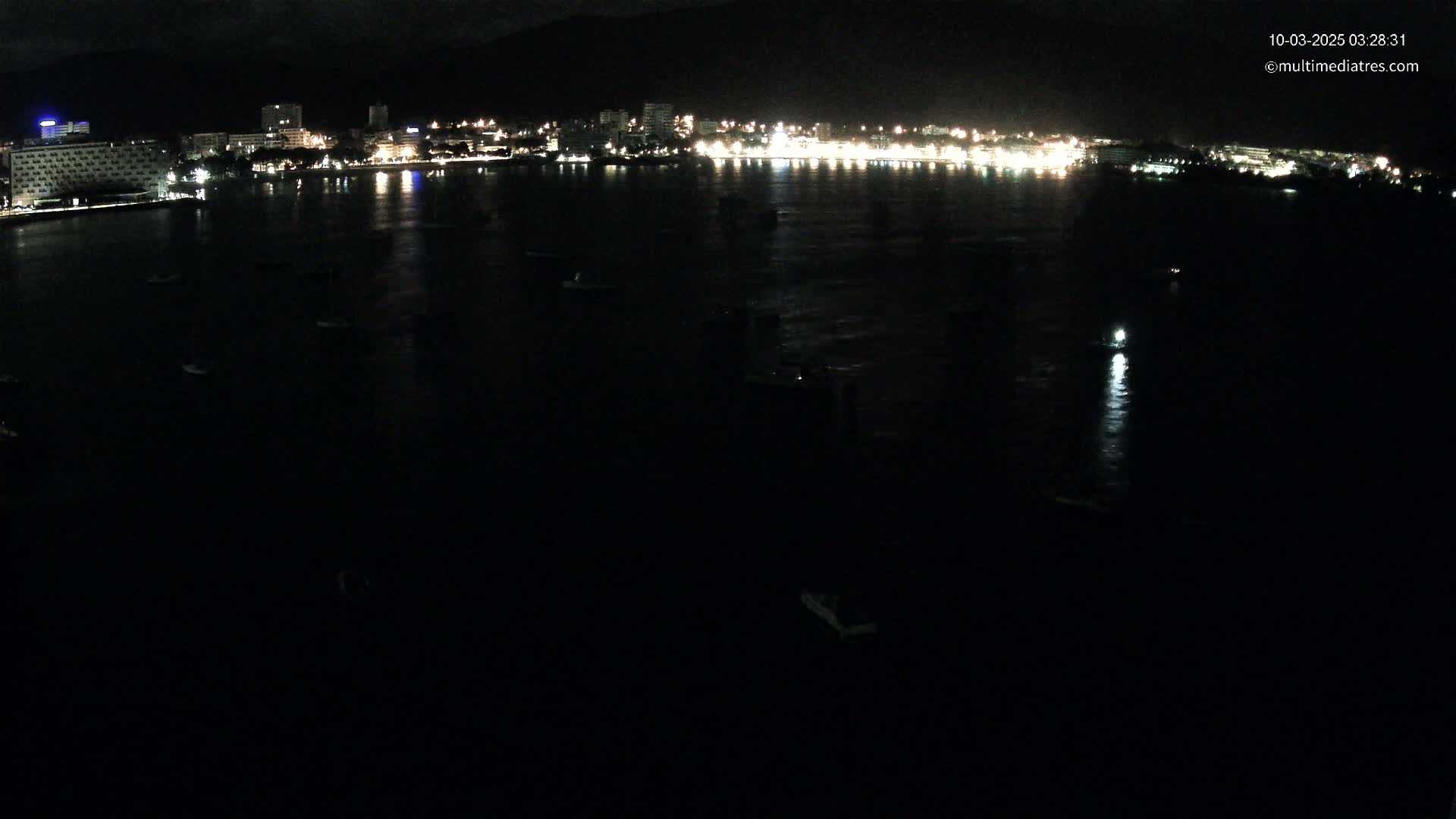 A nighttime, aerial view shows a dark body of water with numerous small boats and a brightly lit city skyline in the distance.