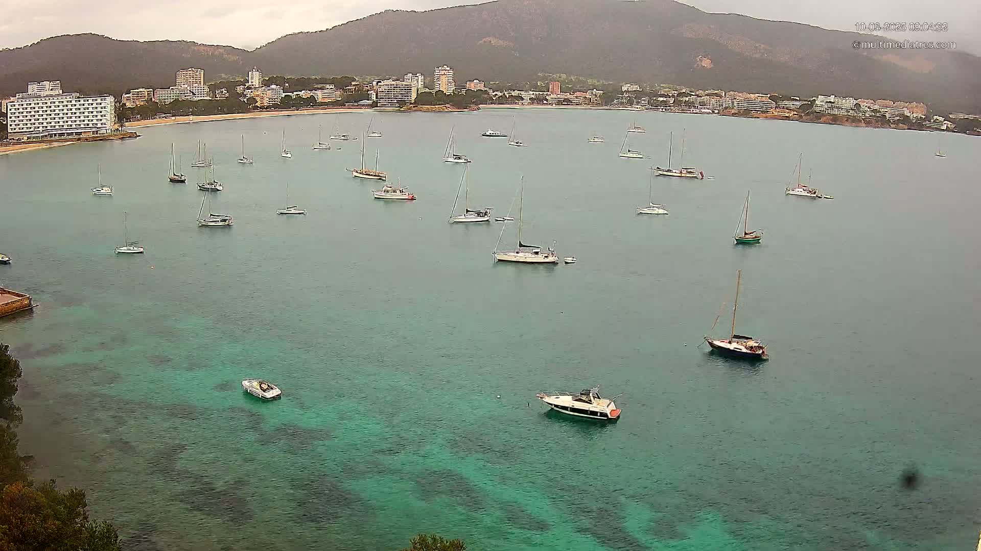 Numerous sailboats and motorboats are anchored in a calm, teal bay in front of a coastal town nestled beneath a mountain range on an overcast day.