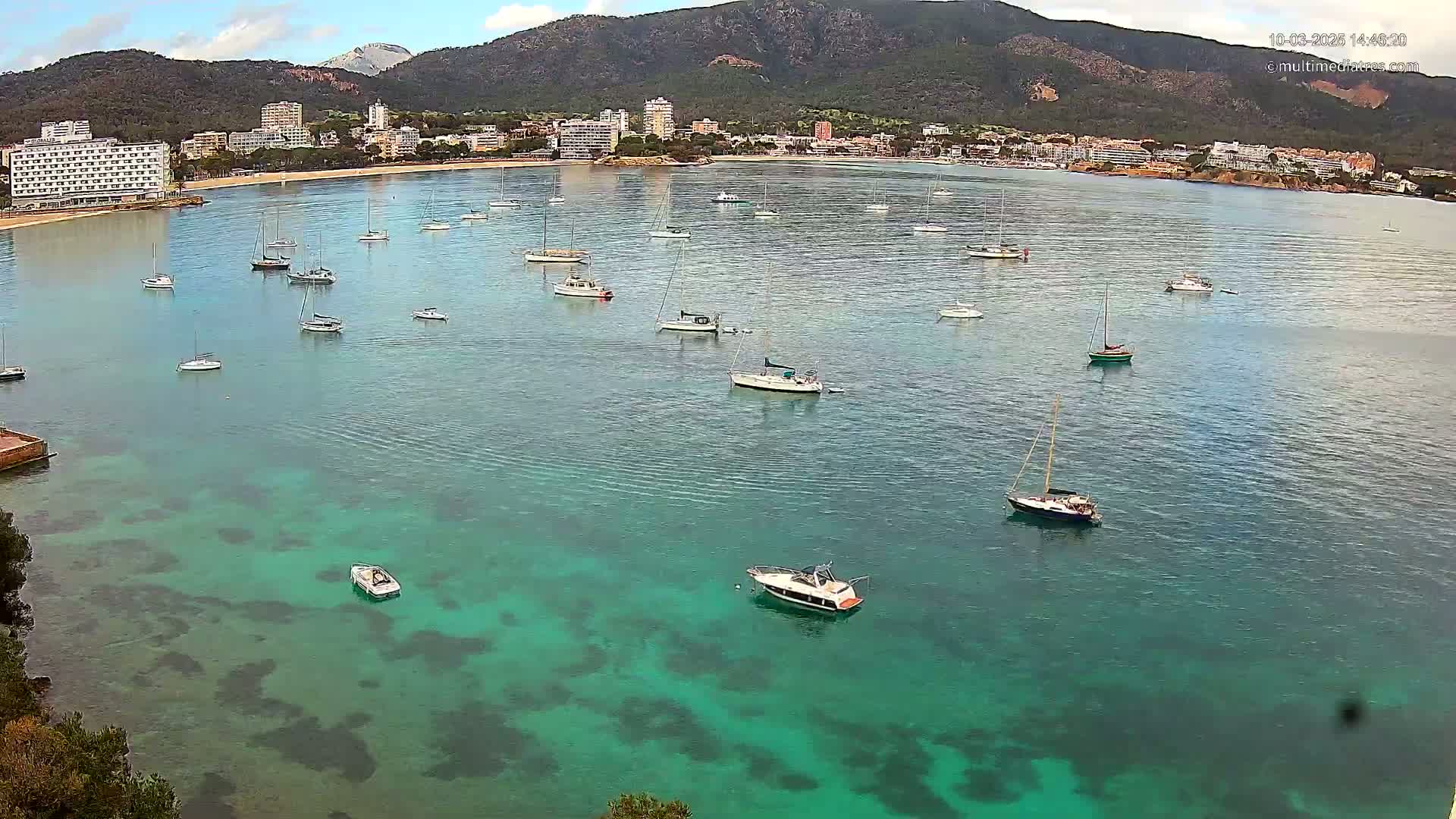 A calm bay with numerous sailboats and motorboats is seen from an aerial perspective, with a town and mountains in the background under a partly cloudy sky.