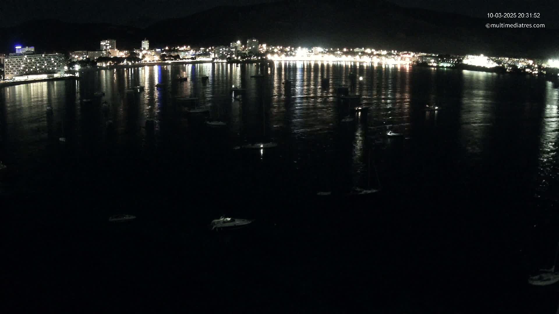 A nighttime view of a calm body of water with numerous boats moored, reflecting the lights of a city on the opposite shore.