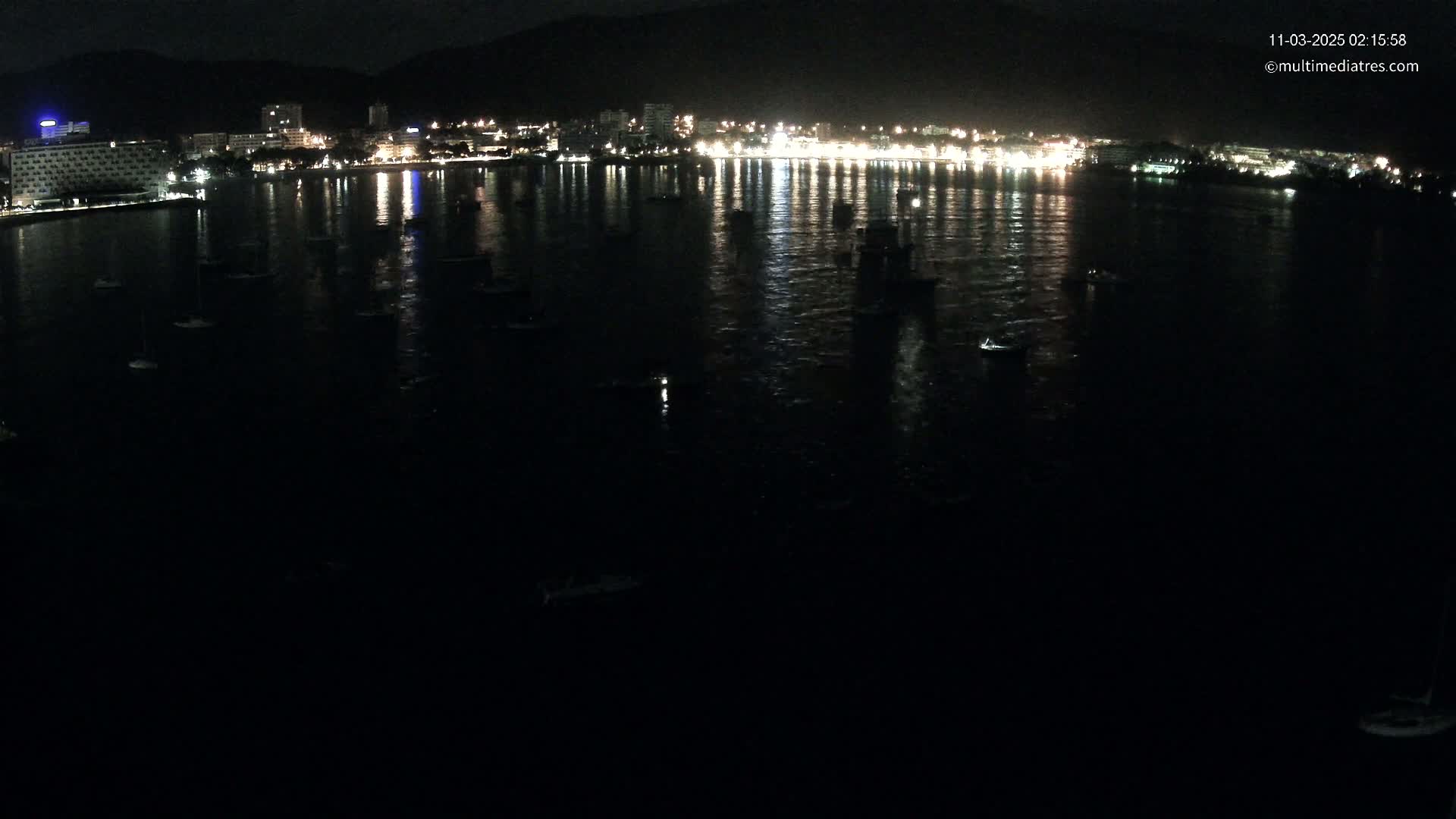 At night, numerous boats are moored in a dark body of water in front of a brightly lit waterfront town nestled against dark mountains.