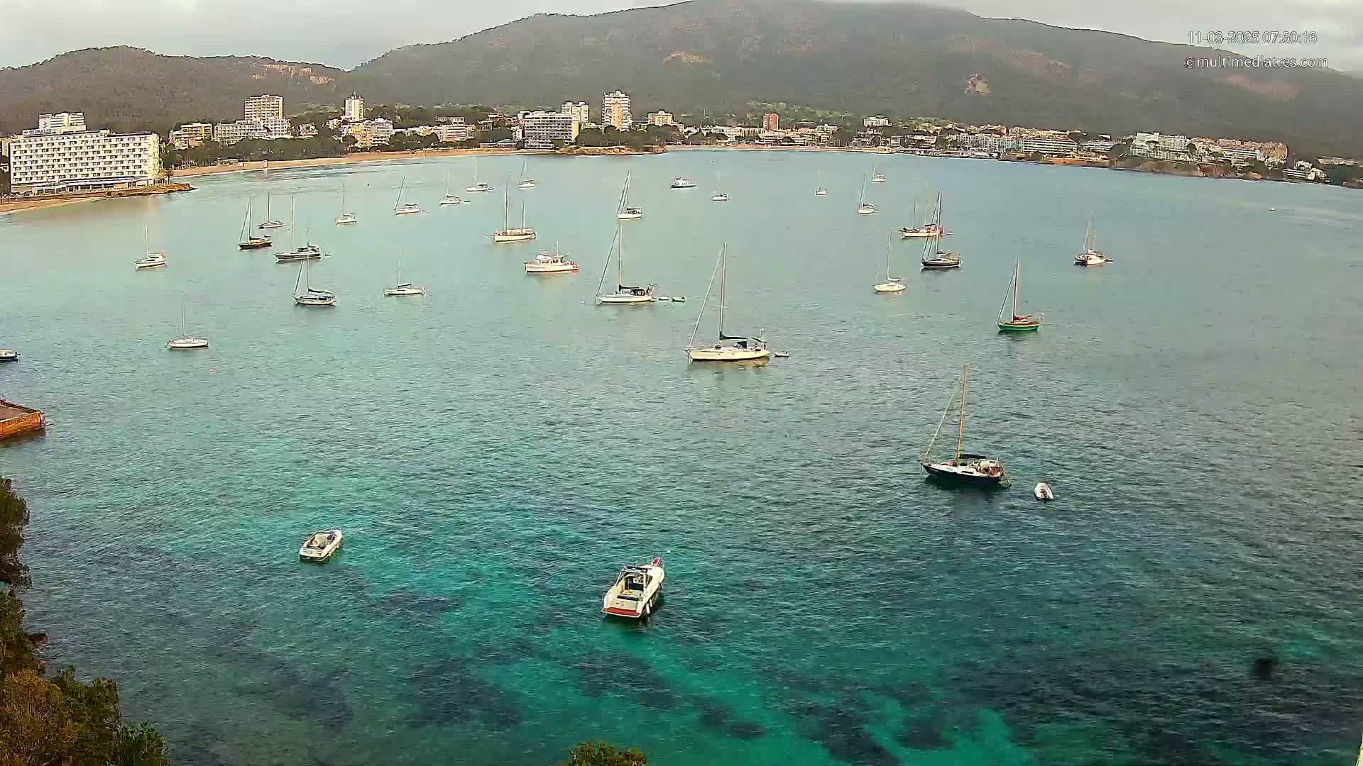 Numerous sailboats and motorboats are anchored in a calm, clear turquoise bay, with a town and mountains visible in the background under an overcast sky.