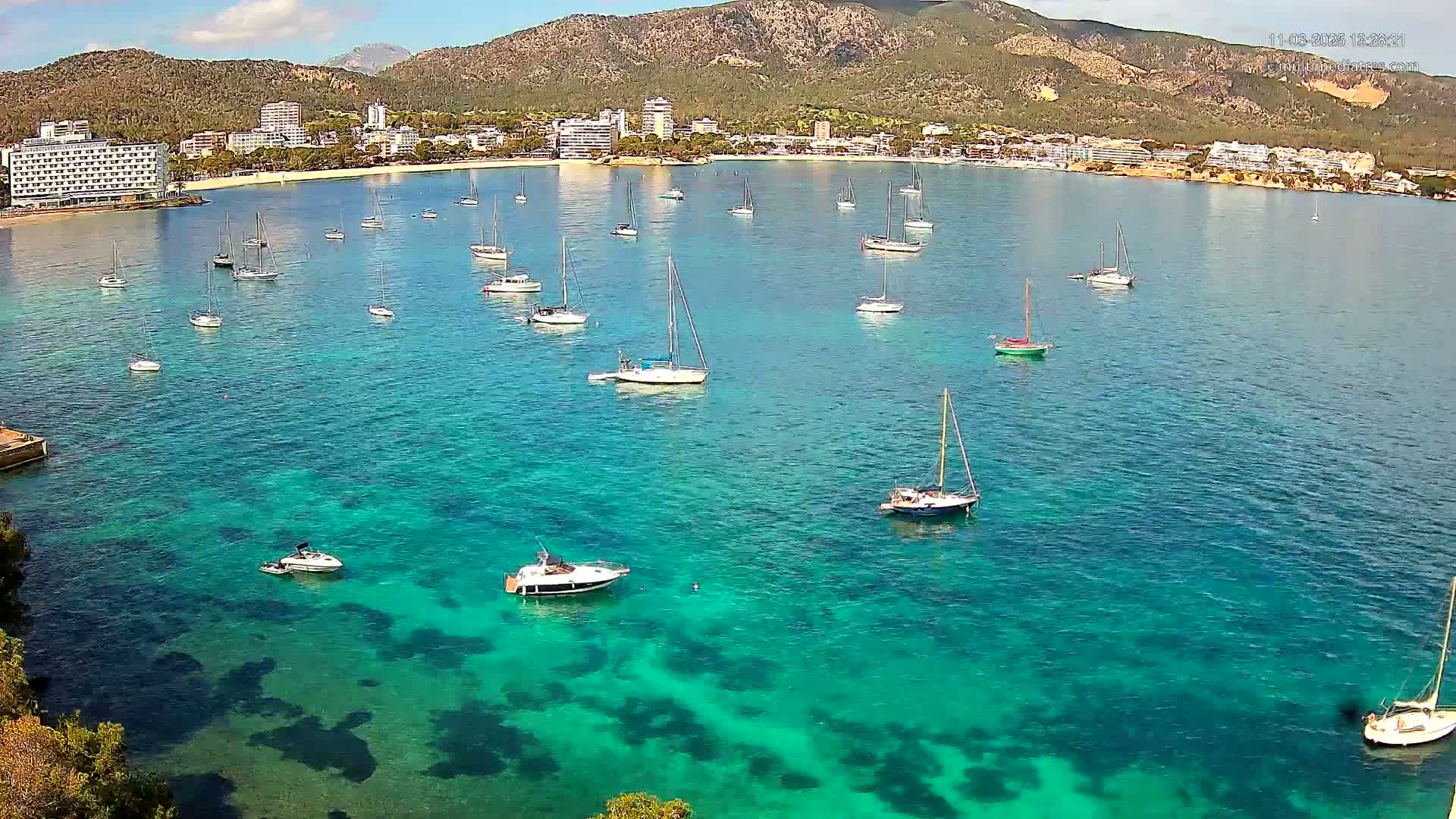 Numerous sailboats are anchored in a calm, clear, turquoise bay near a shoreline with hotels and a mountain in the background under a sunny sky.