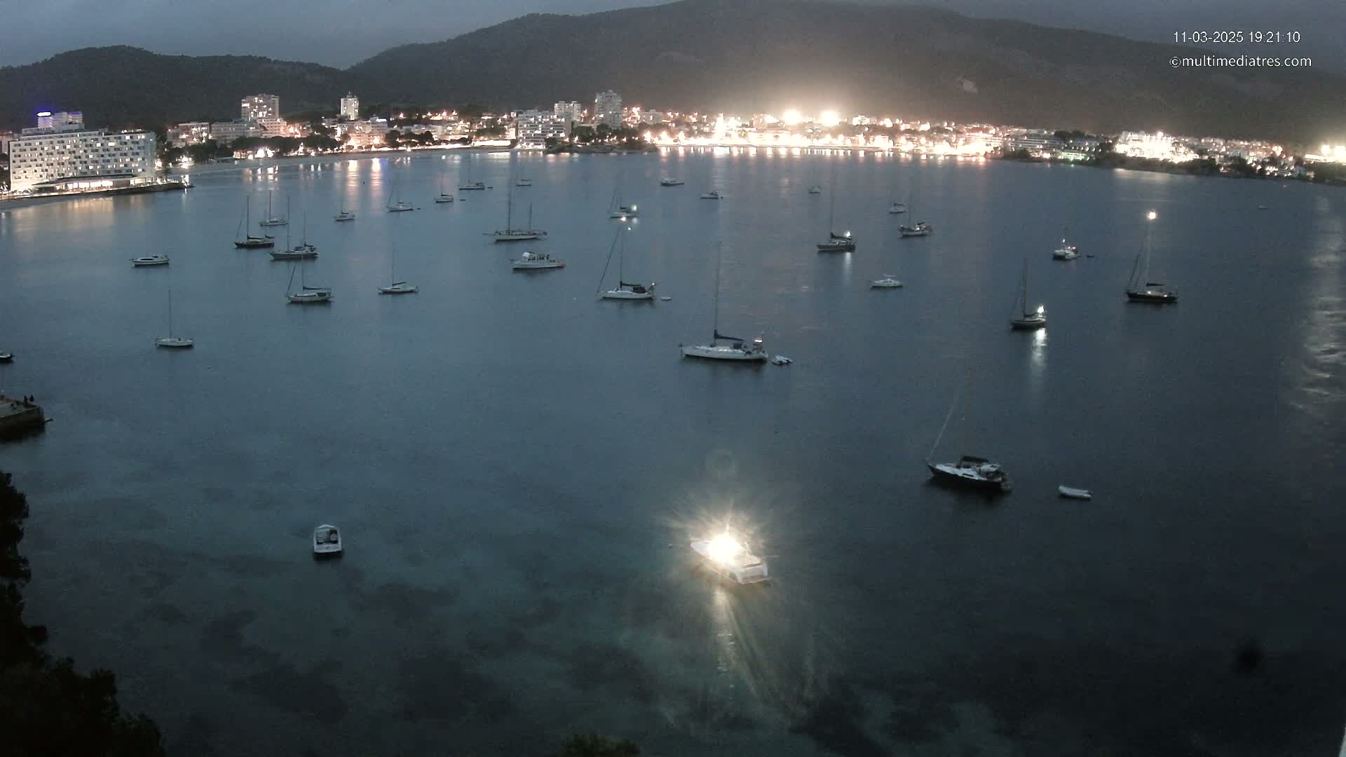 Numerous sailboats are moored in a calm, dark bay at night, in front of a brightly lit town nestled against a mountain.