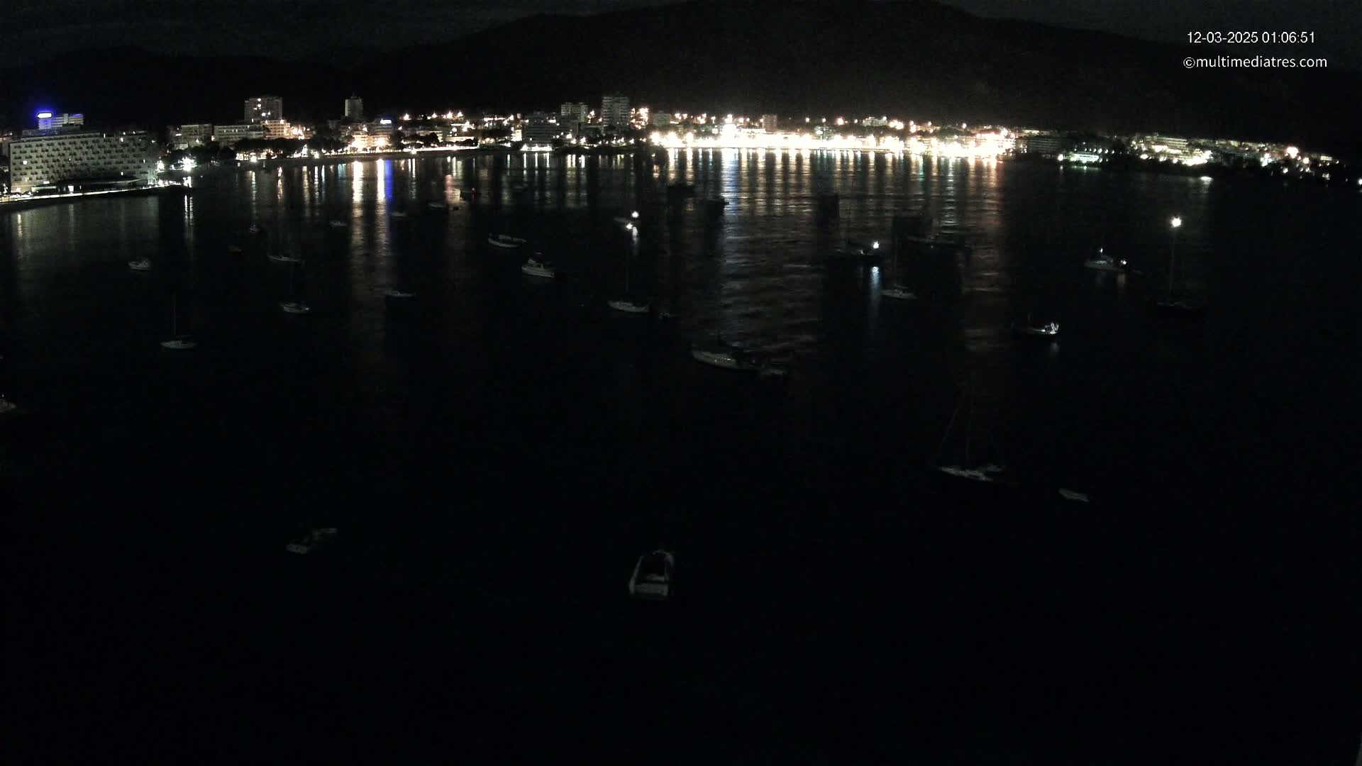 A nighttime view of a city's illuminated waterfront, where numerous boats are moored in a calm body of water.