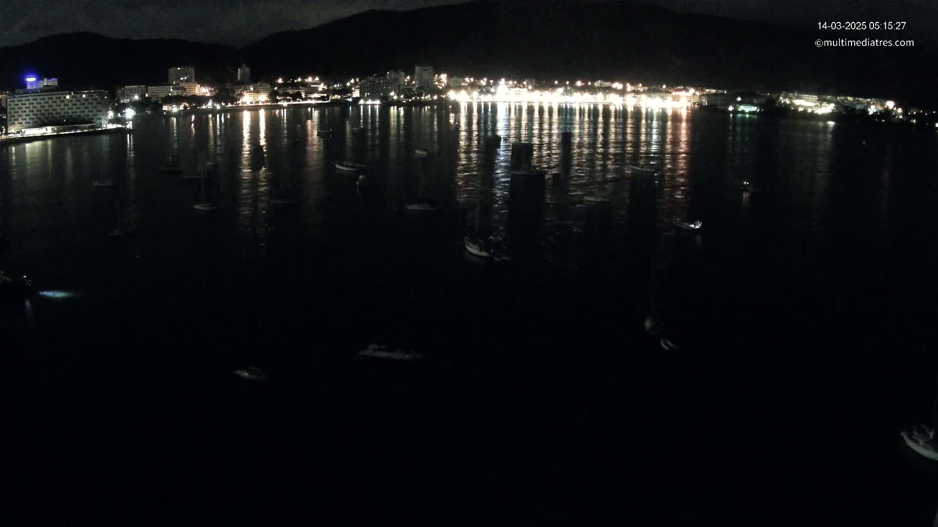A nighttime view of a calm body of water with numerous small boats scattered across its surface, reflecting the lights of a town on the far shore.