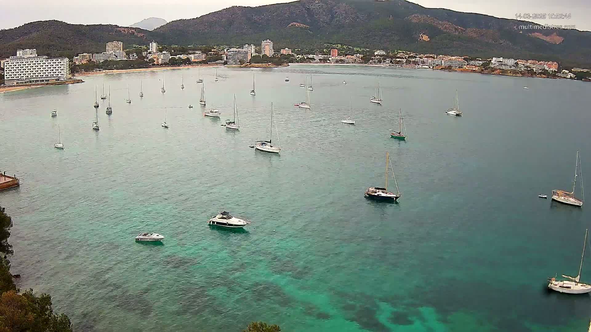 Numerous sailboats and motorboats are anchored in a calm, teal bay near a shoreline with hotels and mountains under an overcast sky.