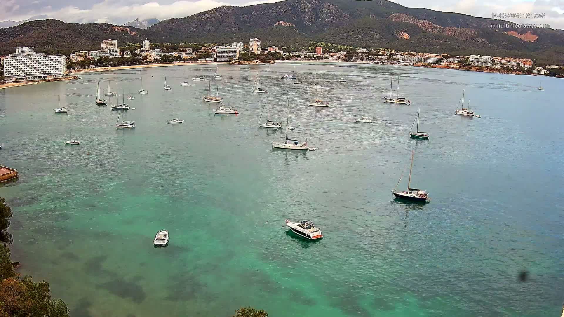 Numerous sailboats and motorboats are anchored in a calm, clear, teal bay near a shoreline with hotels and a mountainous backdrop under an overcast sky.