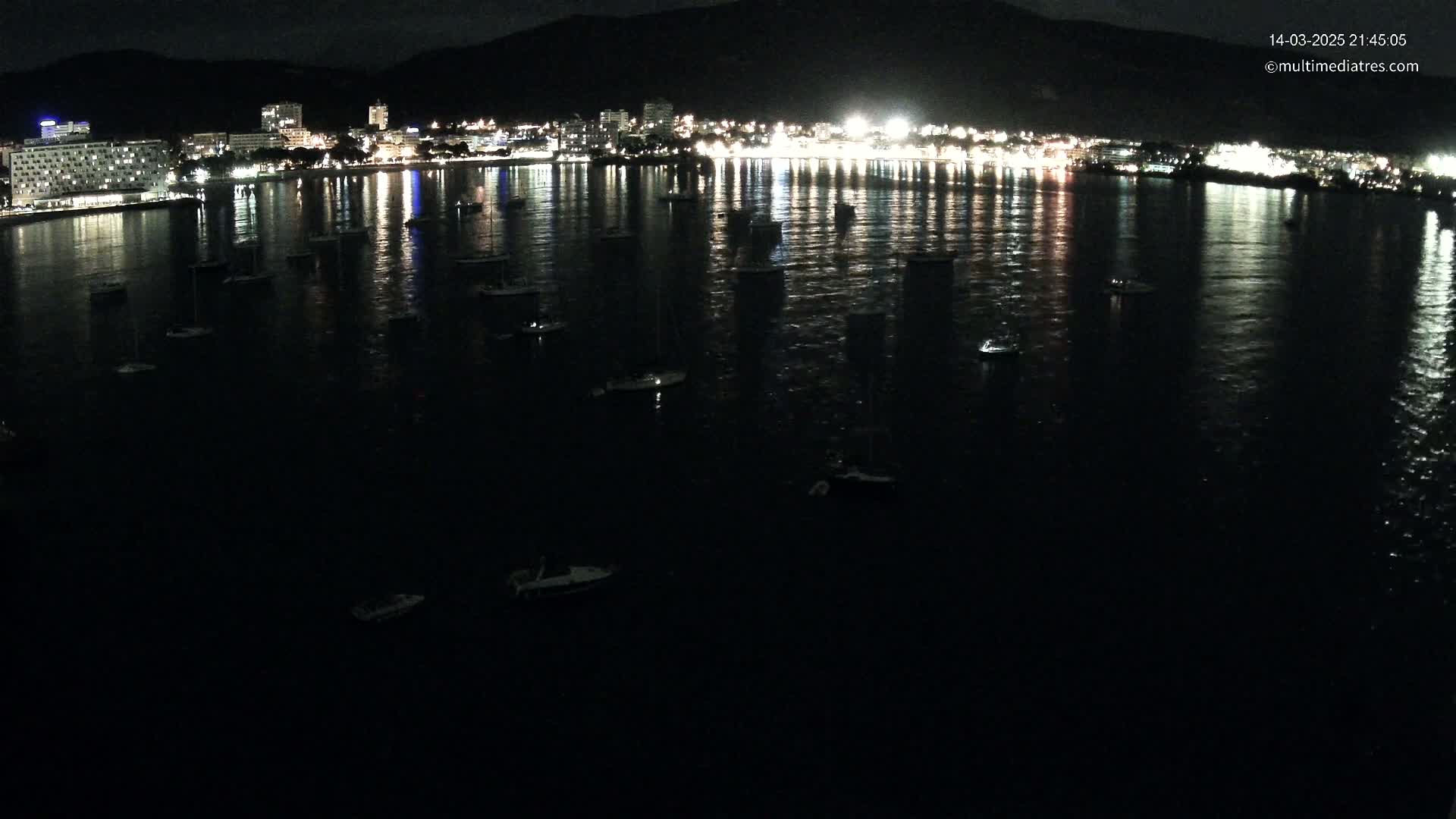 A nighttime view of a calm bay with numerous small boats, reflecting the lights of a city on the shore under a dark sky.