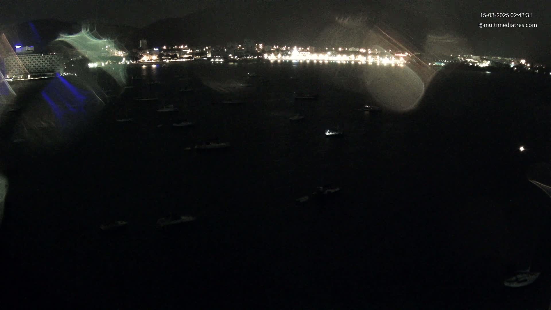 Numerous boats are moored in dark water at night, in front of a city illuminated by lights.