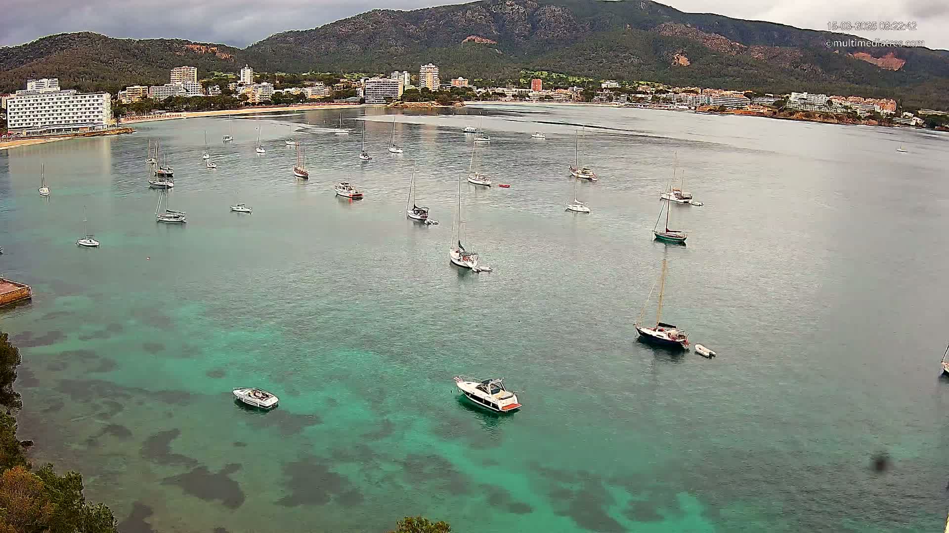 A calm, turquoise bay with numerous sailboats and motorboats anchored near a coastline featuring hotels and a mountainous backdrop under an overcast sky.