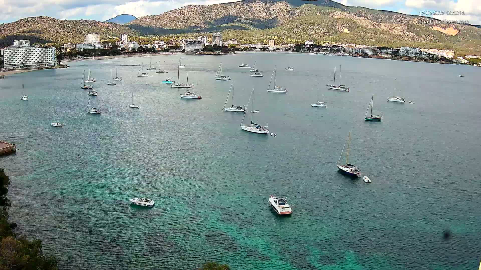 Numerous sailboats and motorboats are anchored in a calm, teal-colored bay, with a mountainous coastline and buildings in the background under a partly cloudy sky.