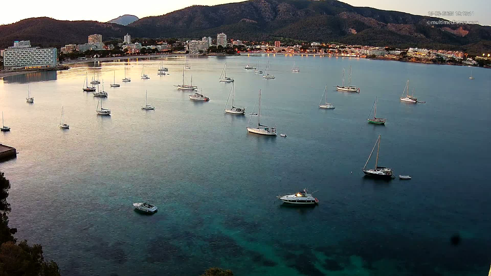 Numerous sailboats are moored in a calm bay near a coastal town at dusk, with mountains visible in the background.