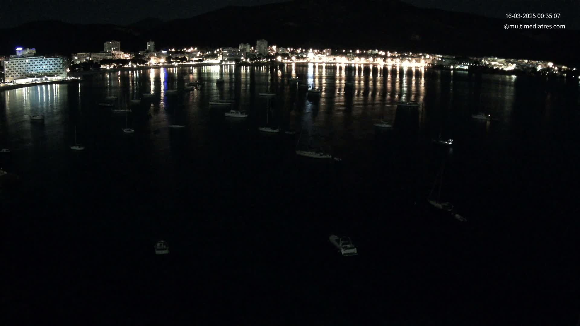 A nighttime aerial view shows a calm body of water with numerous small boats moored, reflecting the lights of a city on the shoreline.