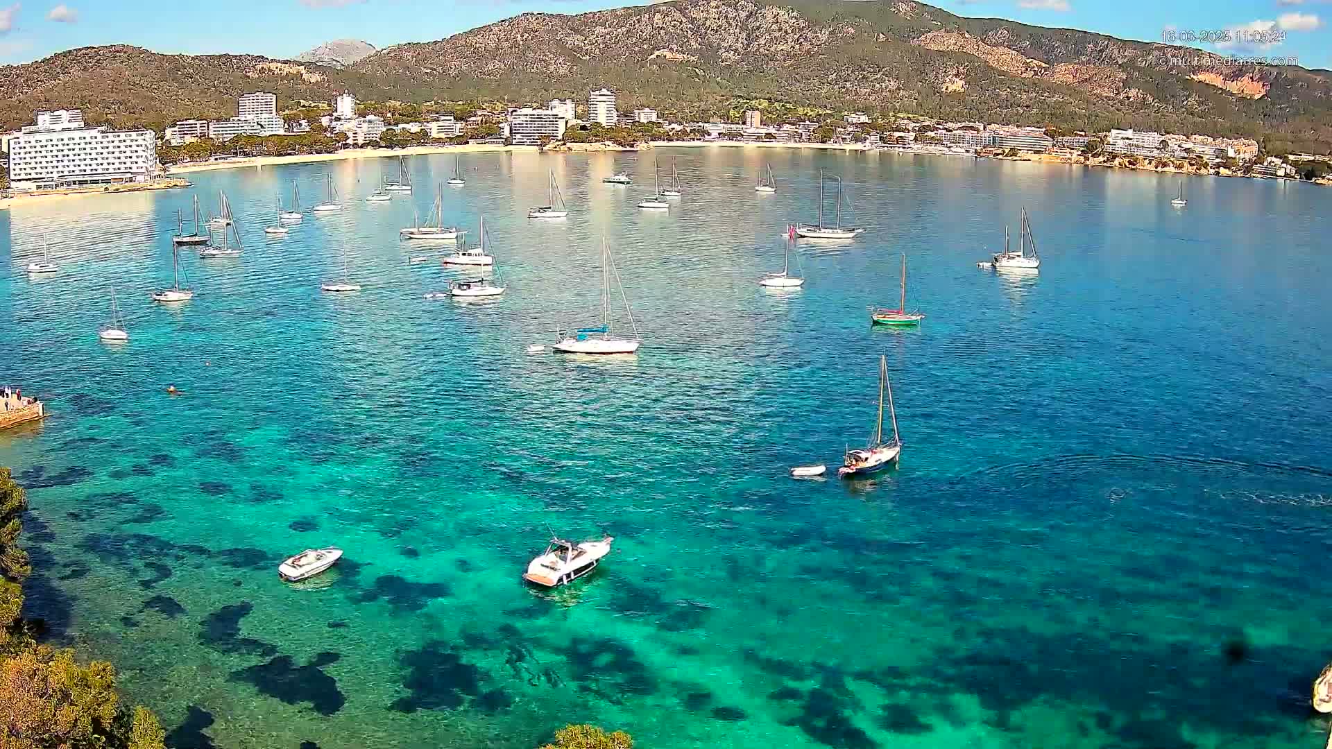 Numerous sailboats and motorboats are anchored in a calm, clear, turquoise bay, near a beach and buildings nestled against a mountain under a sunny sky.