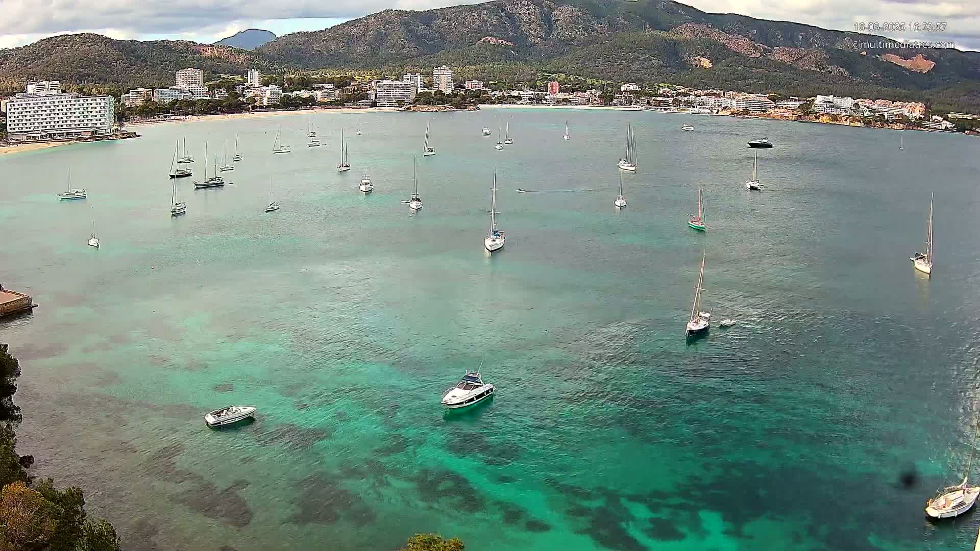 Numerous sailboats and motorboats are scattered across a calm, teal-colored bay, with a shoreline of buildings and a mountainous backdrop under a partly cloudy sky.