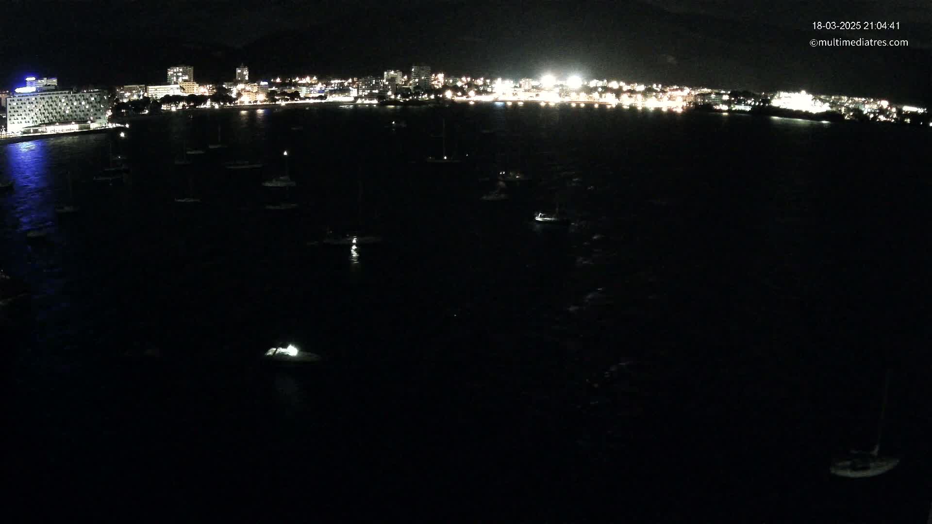 A nighttime aerial view shows a dark body of water with numerous small boats at anchor in front of a brightly lit city.