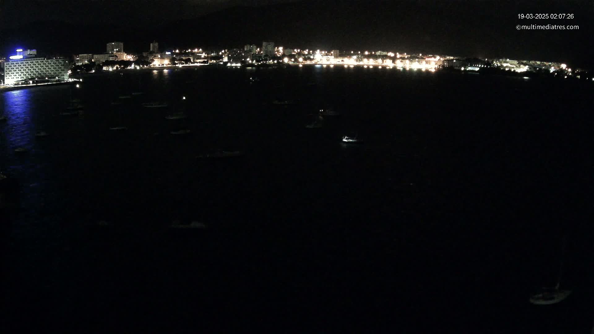 A nighttime, dark-water bay with numerous small boats scattered across its surface, and a brightly lit city skyline in the background.