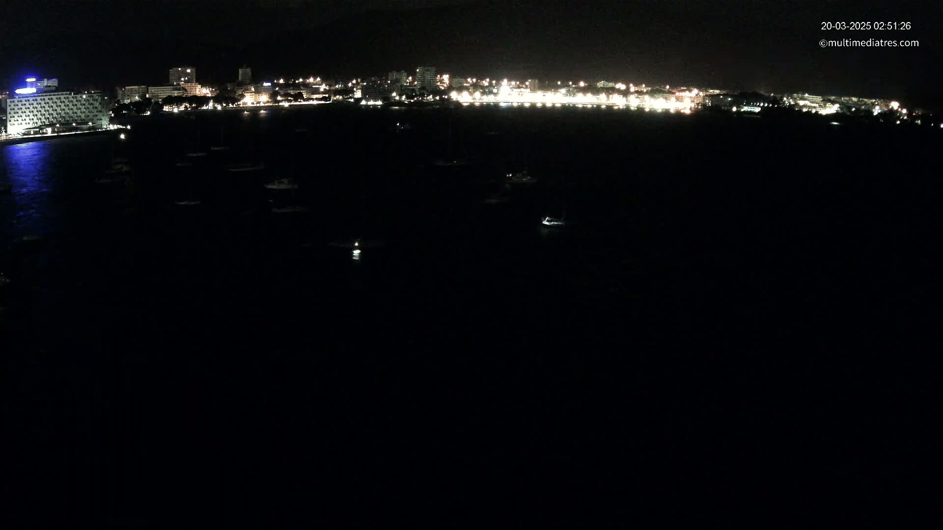 A nighttime view of a city's illuminated waterfront at night, showing many small boats in dark water.