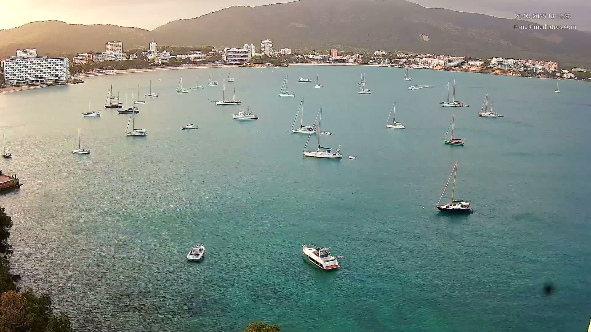 Numerous sailboats and motorboats are anchored in a calm, teal-colored bay, with a town and mountains visible in the background under a somewhat overcast sky.