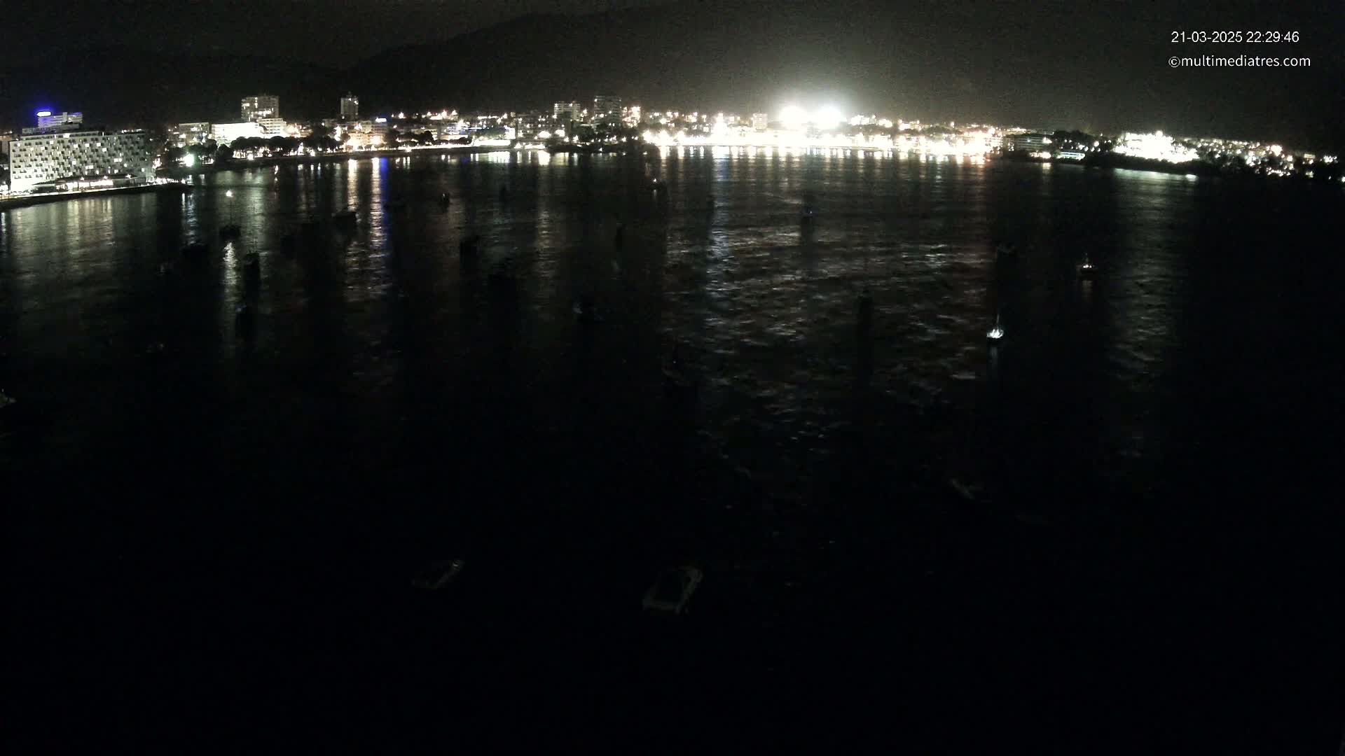 A nighttime view shows a dark body of water with several small boats, reflecting the lights of a city on the shore.