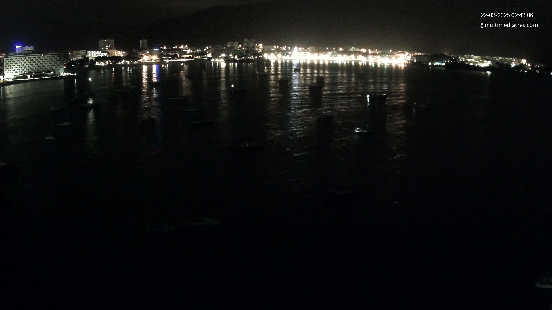 A nighttime view of a calm body of water with numerous small boats at rest, reflecting the lights of a city on the shore.