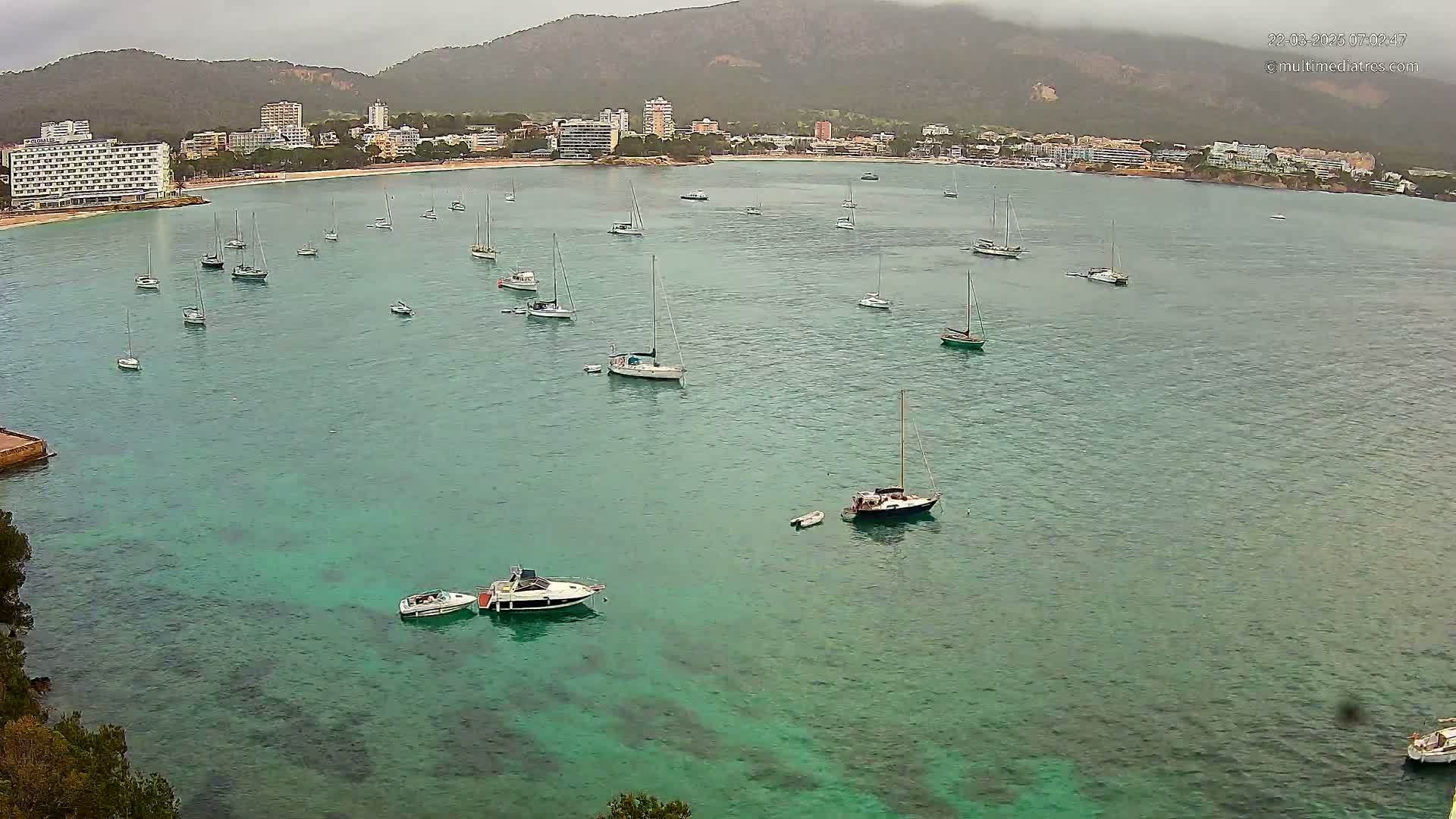 A calm, overcast day shows numerous sailboats and motorboats anchored in a teal bay near a shoreline of hotels and low-lying buildings backed by mountains.