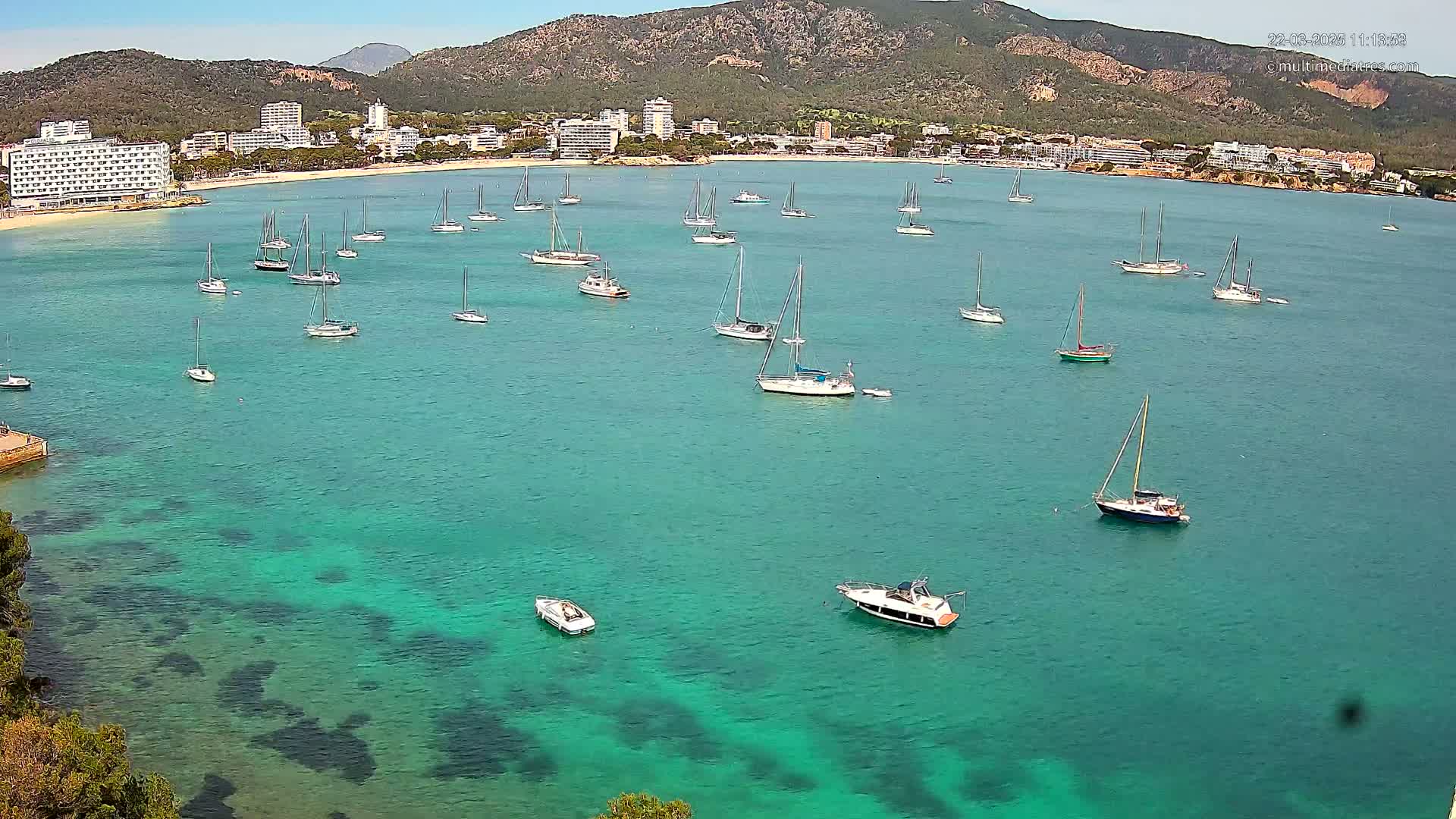 Numerous sailboats and motorboats are anchored in a calm, clear, teal-colored bay, with a mountainous coastline and buildings in the background under a sunny sky.